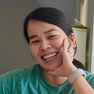 A woman with black hair smiling and resting her face on her hand in a casual setting.