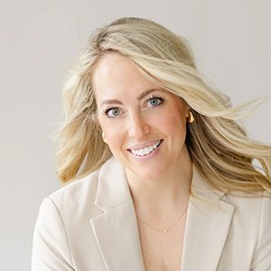 Portrait of a smiling woman with long blonde hair, wearing a light-colored blazer against a neutral background.