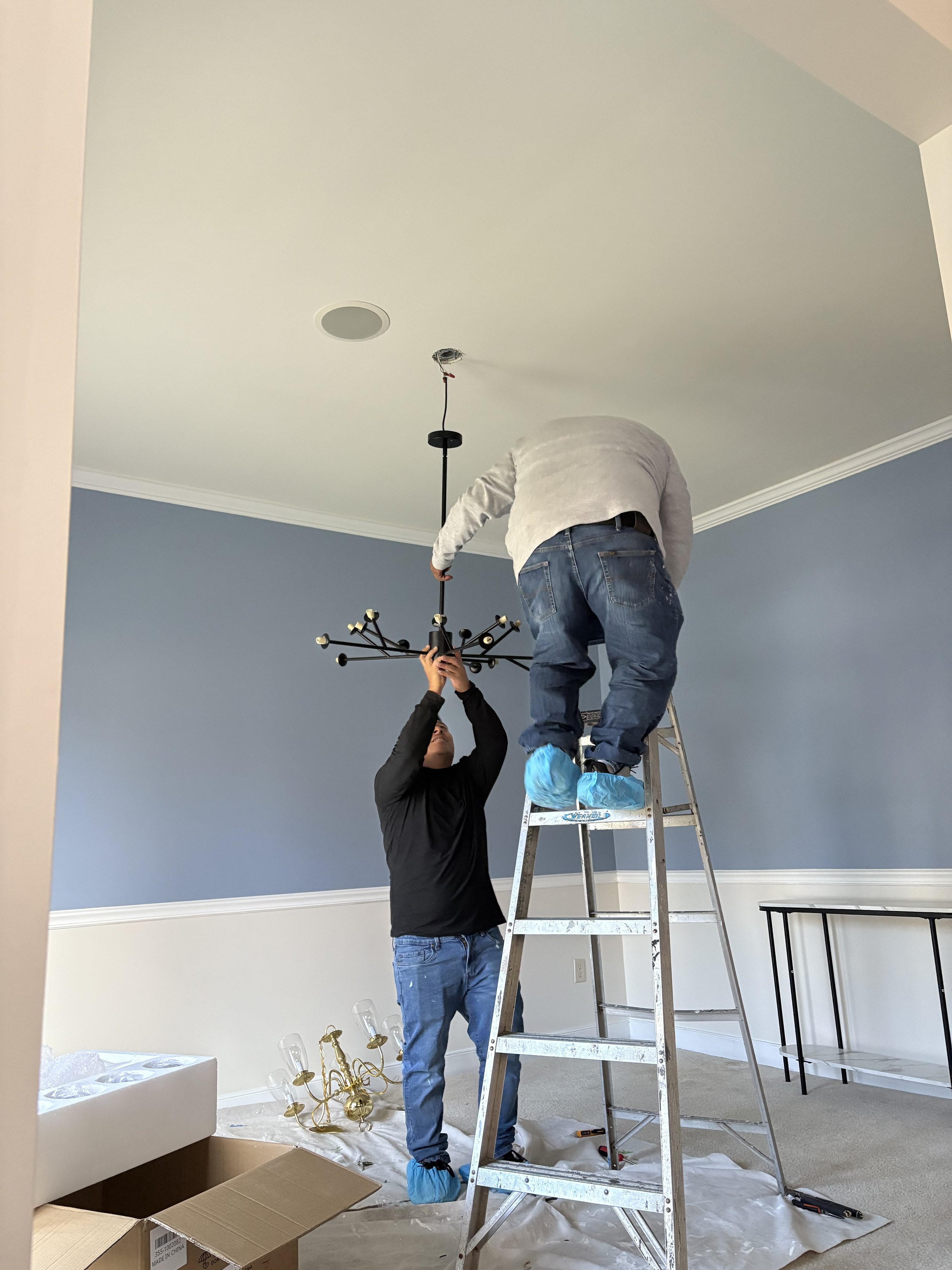 Two people are installing a chandelier in a room with blue walls. One person is standing on a ladder, while the other is holding the light fixture. A previously uninstalled chandelier and open box are on the floor. Both individuals are wearing protec