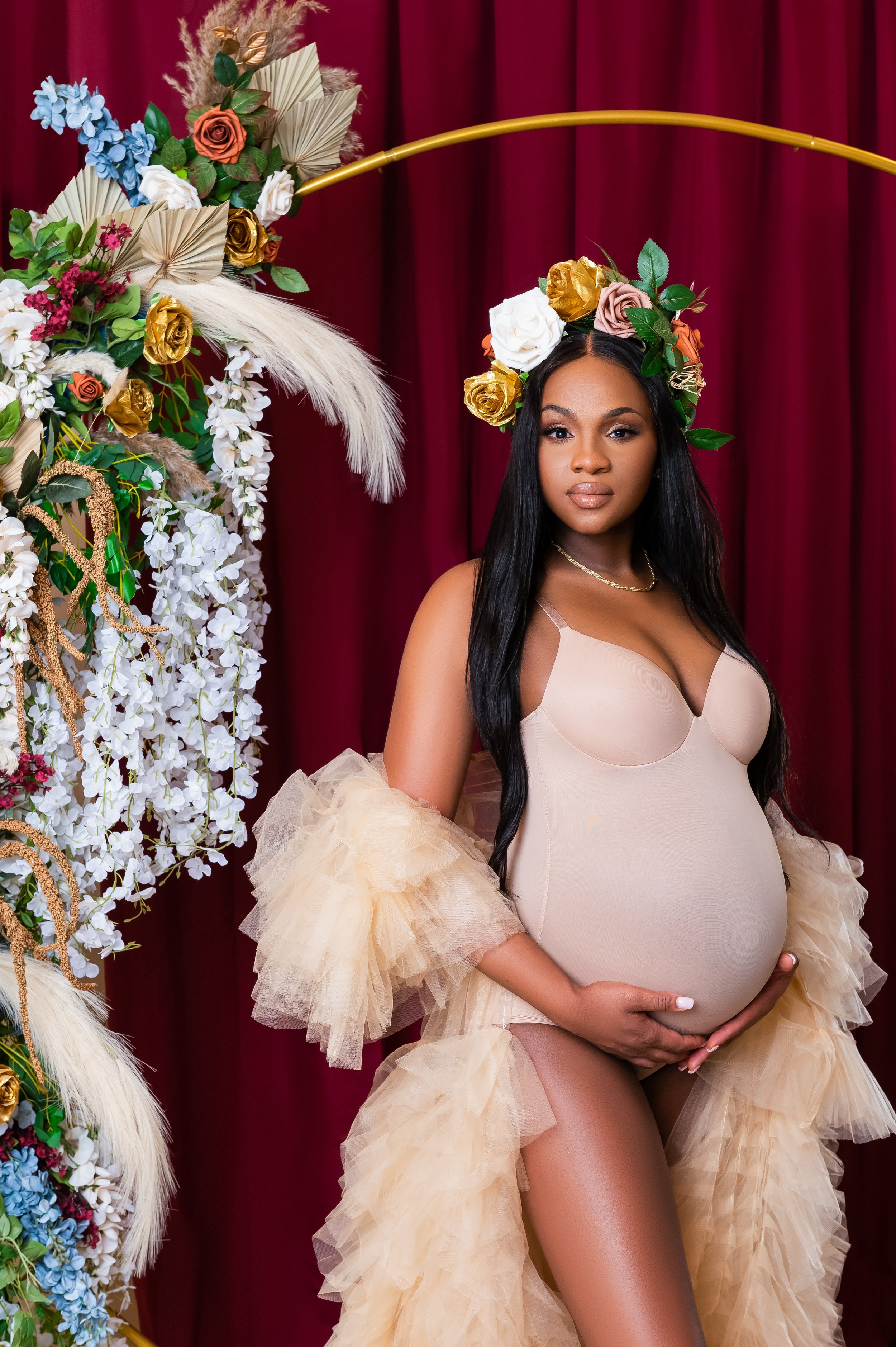 Maternity photoshoot, Pregnant woman in beige dress with floral headdress in front of a red curtain, standing beside a floral arrangement.