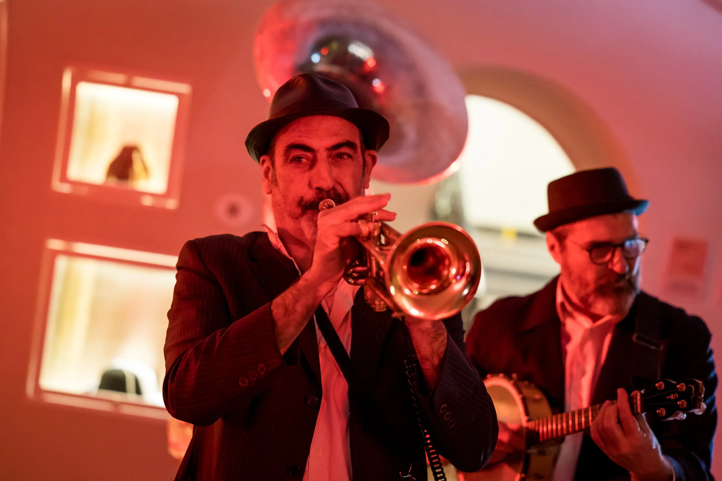 Two men in suits and hats playing musical instruments, one with a trumpet and the other with a guitar, in a dimly lit room with colorful lighting.