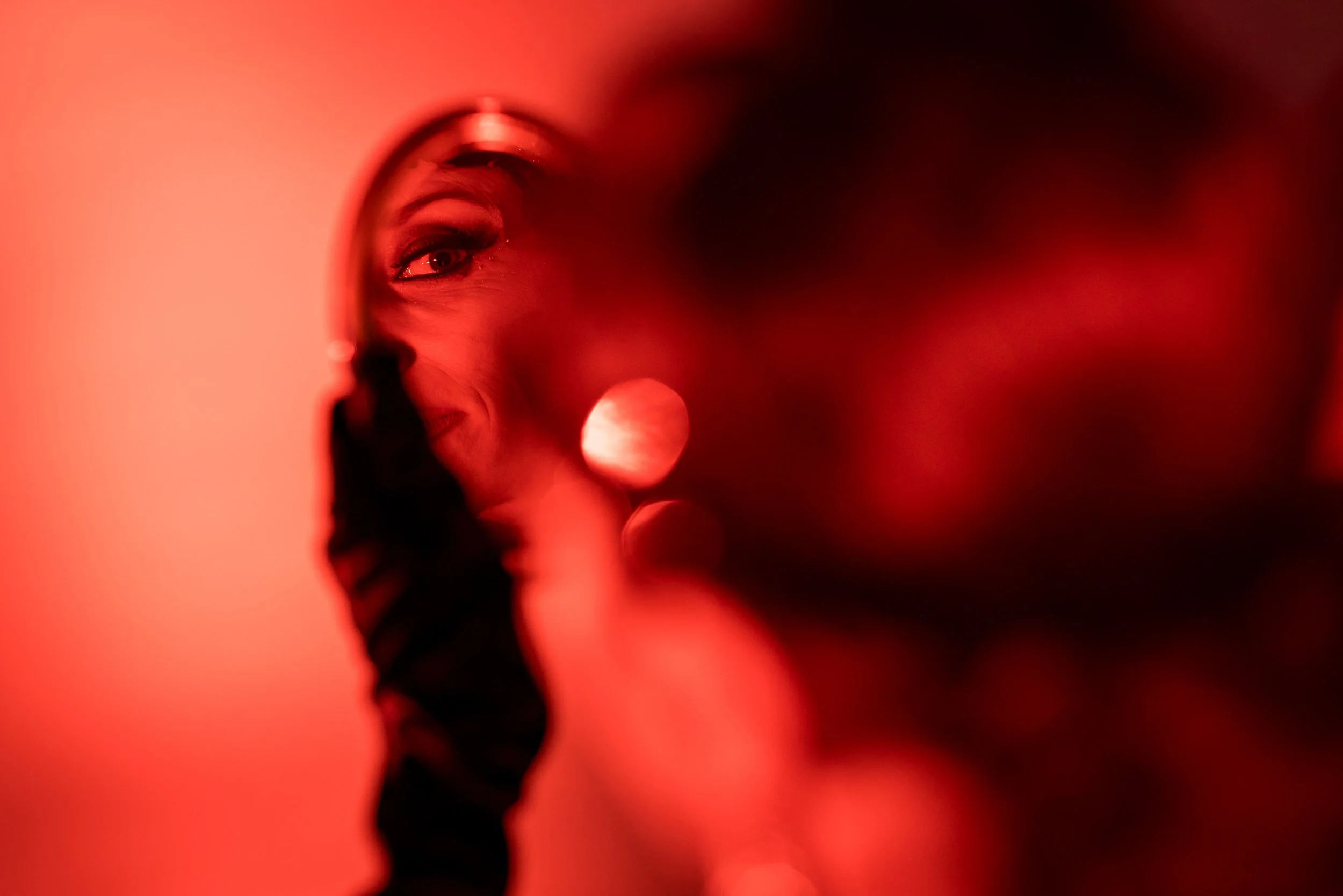 A person looking into a small handheld mirror, with the reflection showing their eyes and part of their face, illuminated by red lighting.