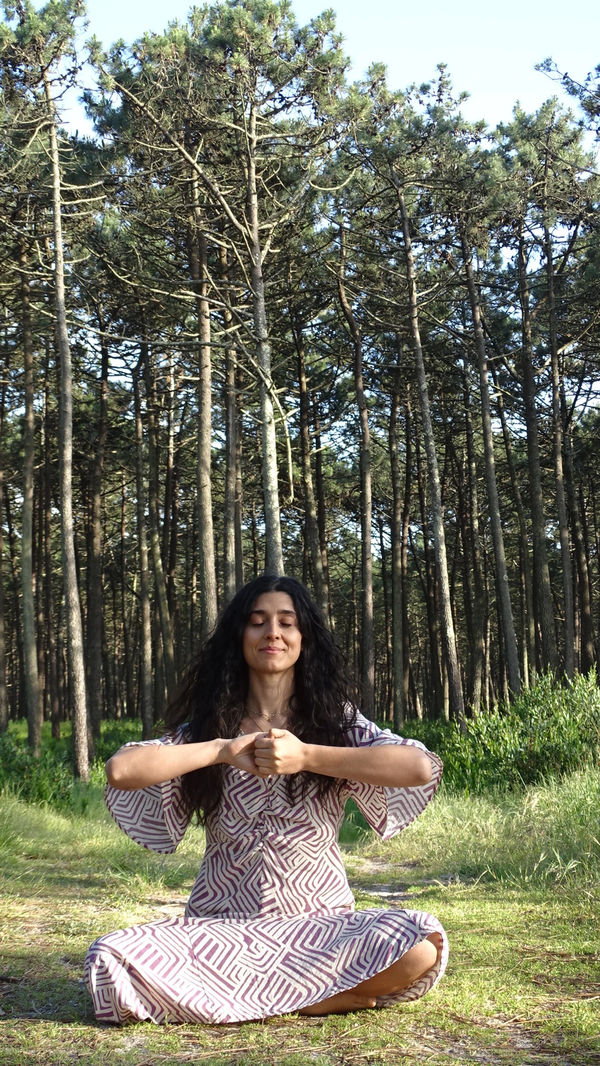 Woman sitting cross-legged on grass in a forest, meditating with eyes closed, in a pink and white patterned dress.
