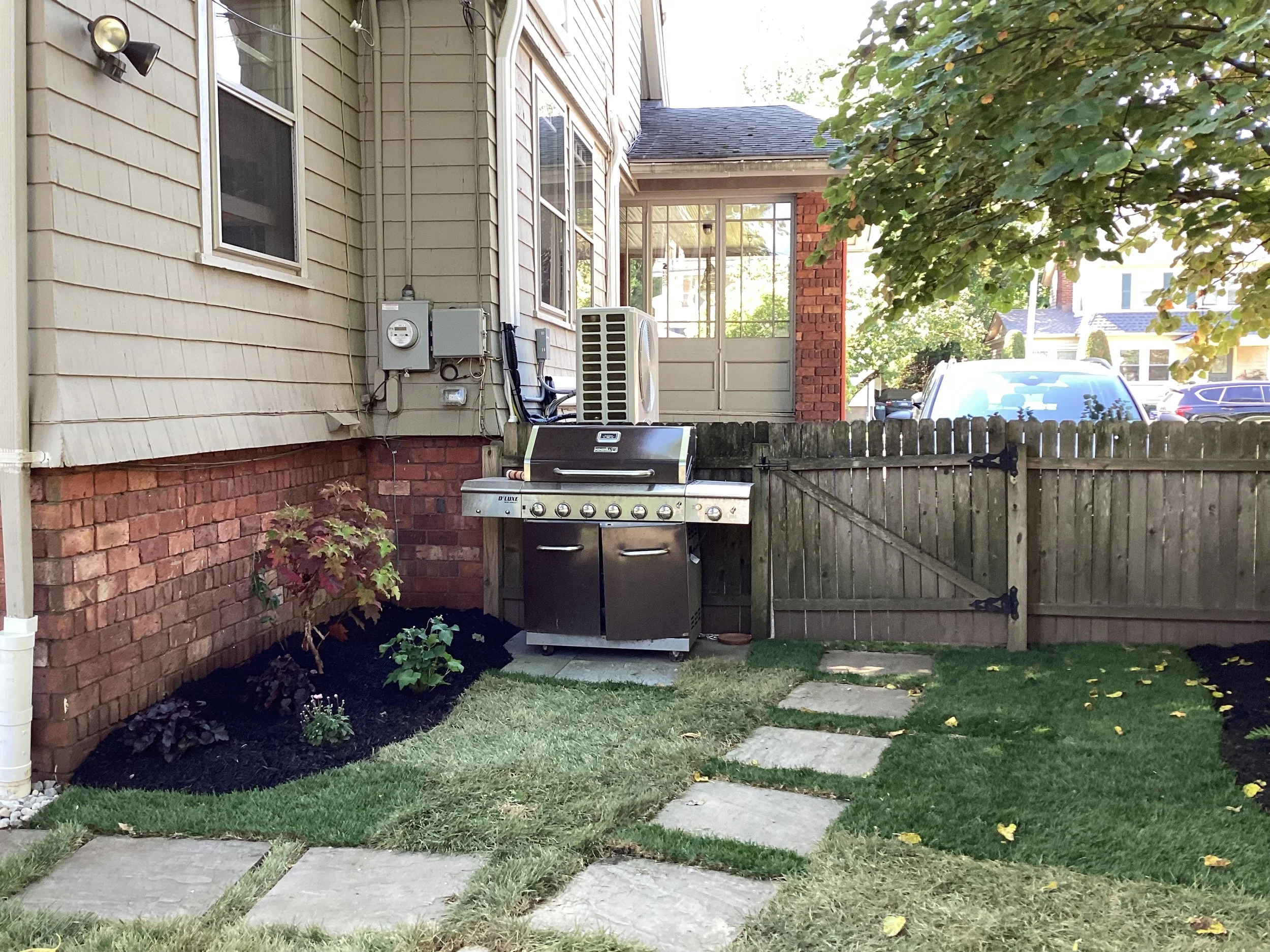 Backyard with a stone pathway, a garden with plants and mulch, a wooden fence with a gate, and a stainless steel barbecue grill next to a house with beige siding and brick foundation. A tree with green leaves extends over part of the yard.