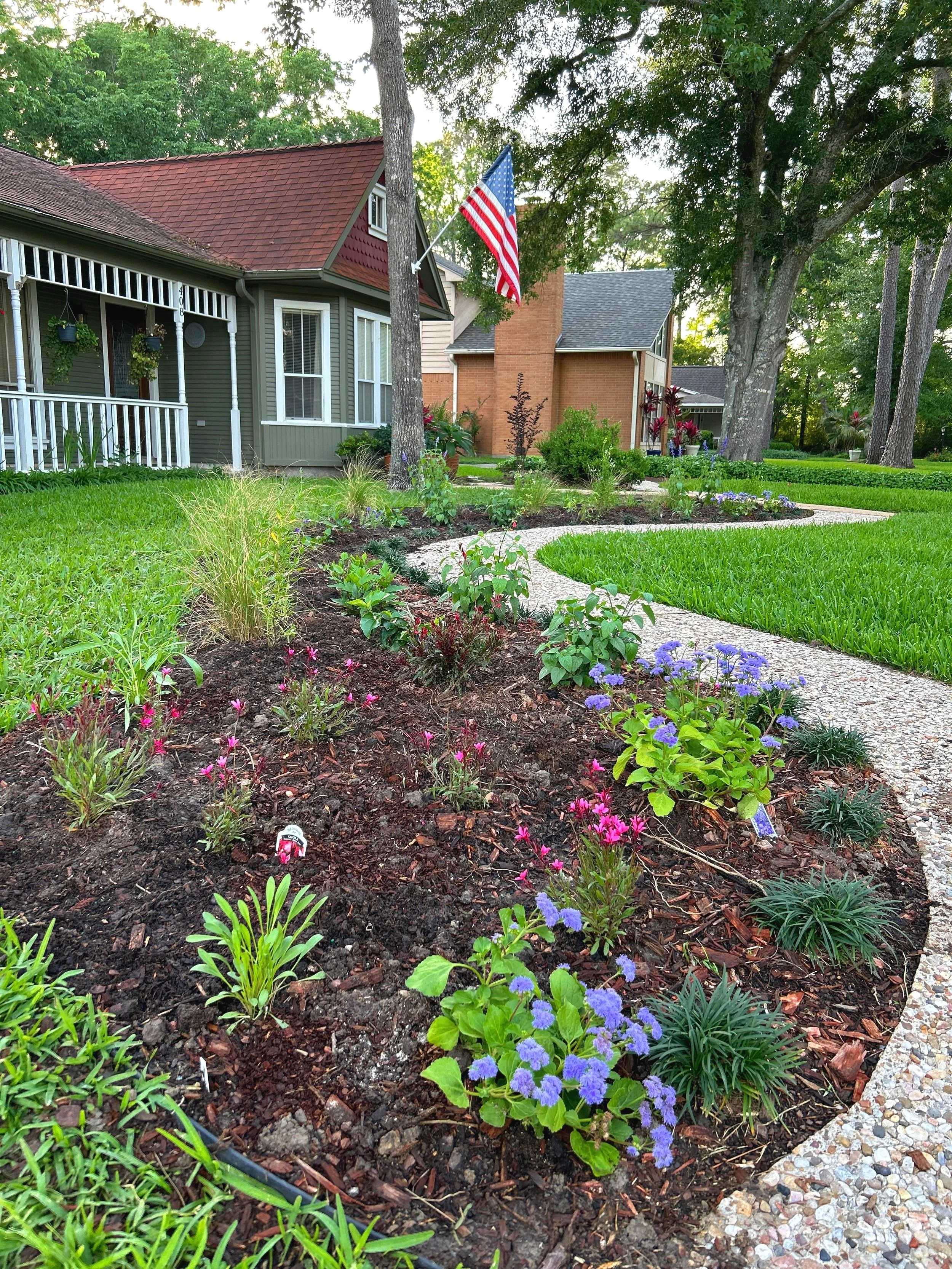 A neatly maintained front yard with a flower bed, a concrete pathway, green grass, trees, and houses in the background. An American flag is mounted on a pole attached to one house.