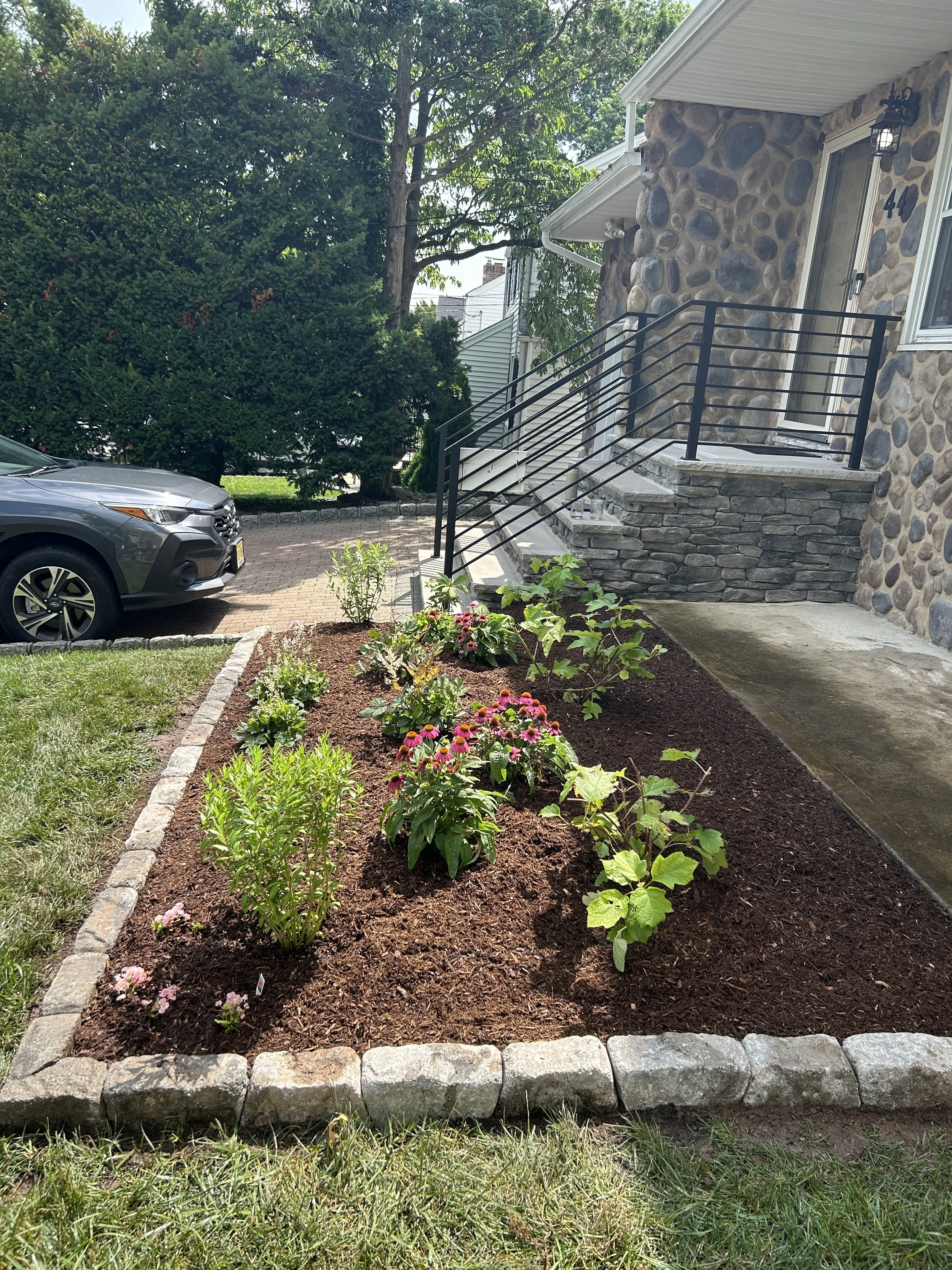 Newly planted flower bed with colorful flowers and green plants outside a stone house, stairs with black metal railing leading to front door, parked car in driveway.