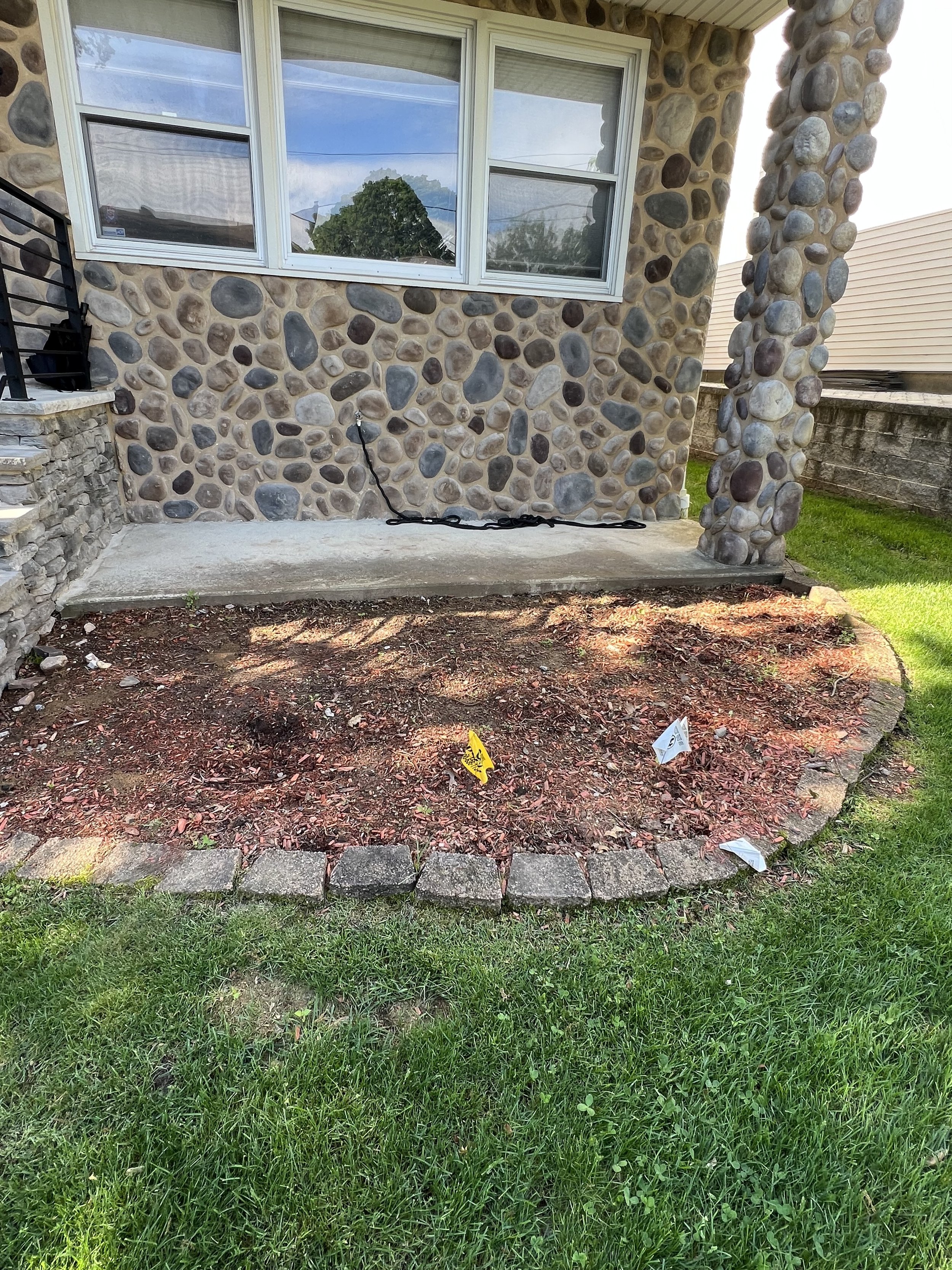 Front yard of a house with a stone exterior and large window. A small garden bed with mulch and bricks borders the grass, a concrete patio in front of the window, and some flags with a garden marker in the mulch.