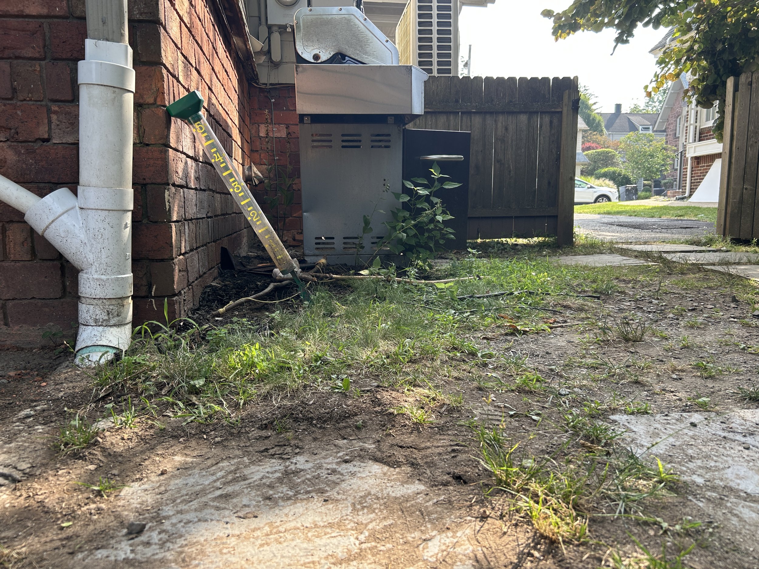 A backyard scene showing a patch of soil with small grass and weeds, a white drainpipe attached to a brick wall, a sprinkler hose on the ground, and an outdoor electrical box nearby. In the background, there is a wooden fence, a sidewalk, a grassy lawn, and a residential neighborhood.