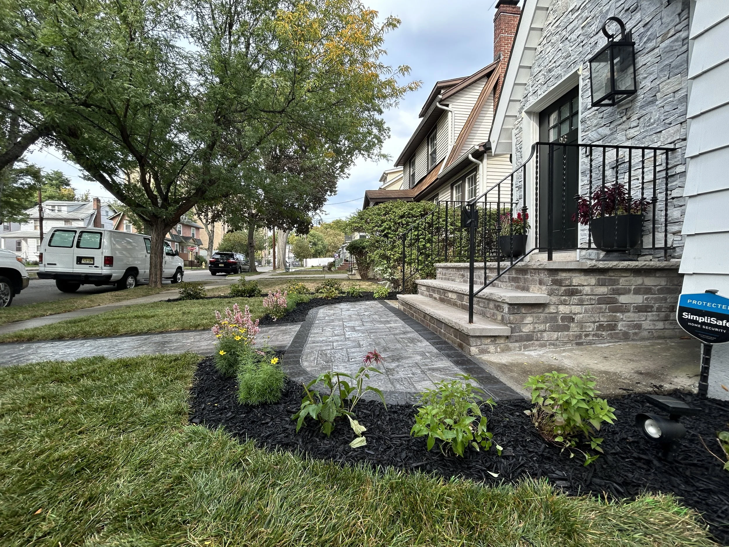 View of a residential front yard with a brick and stone house, including a small porch with steps, potted plants, and black metal railing. A garden with flowers and mulch is in the foreground. There are trees lining the street, parked cars, and neighboring houses visible along the street.