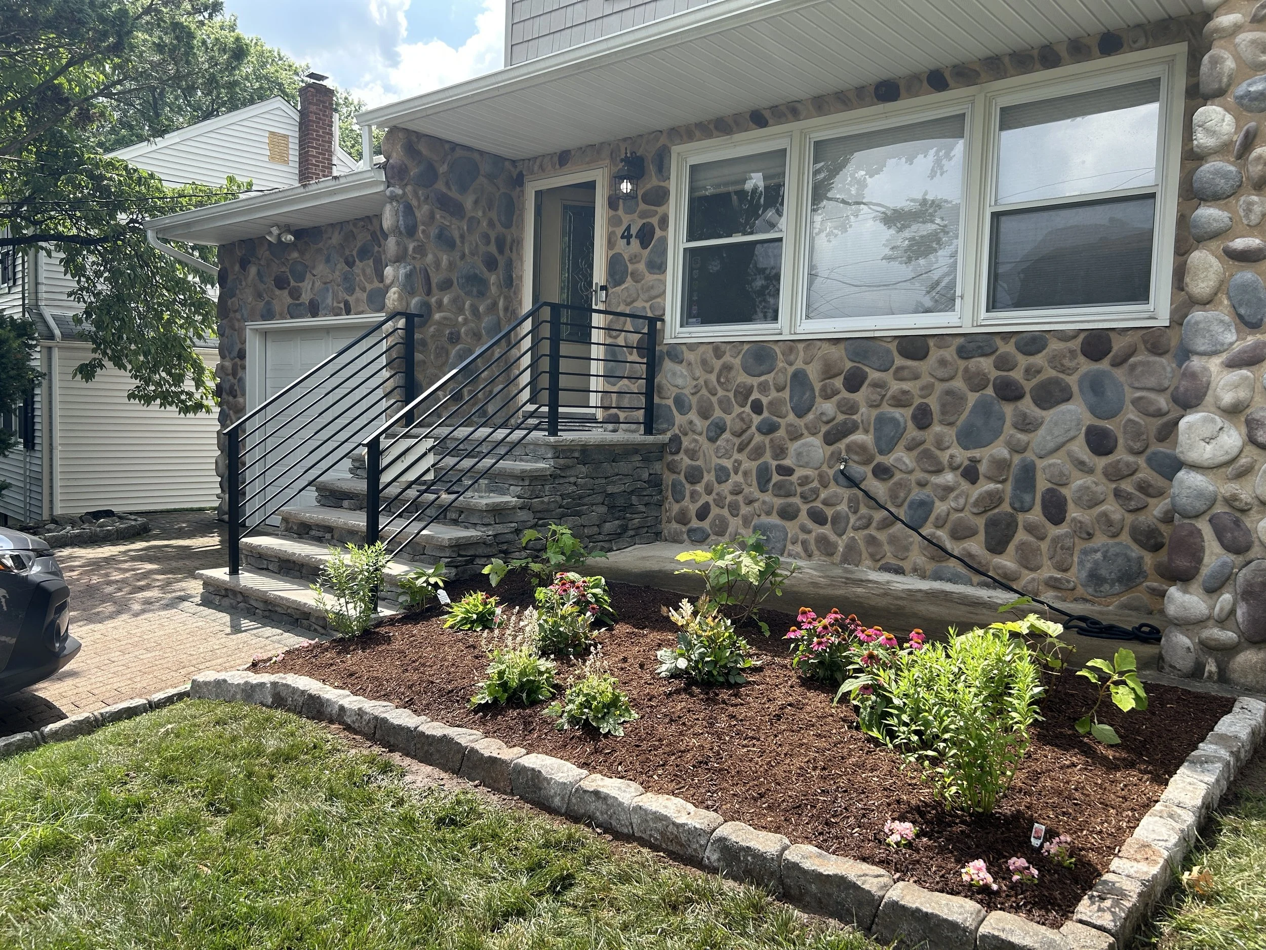 A house with a stone exterior, white window frames, and black metal railings on the front stairs. The house number is 40, and there are flower beds with colorful flowers in front of the house, and a driveway to the side.