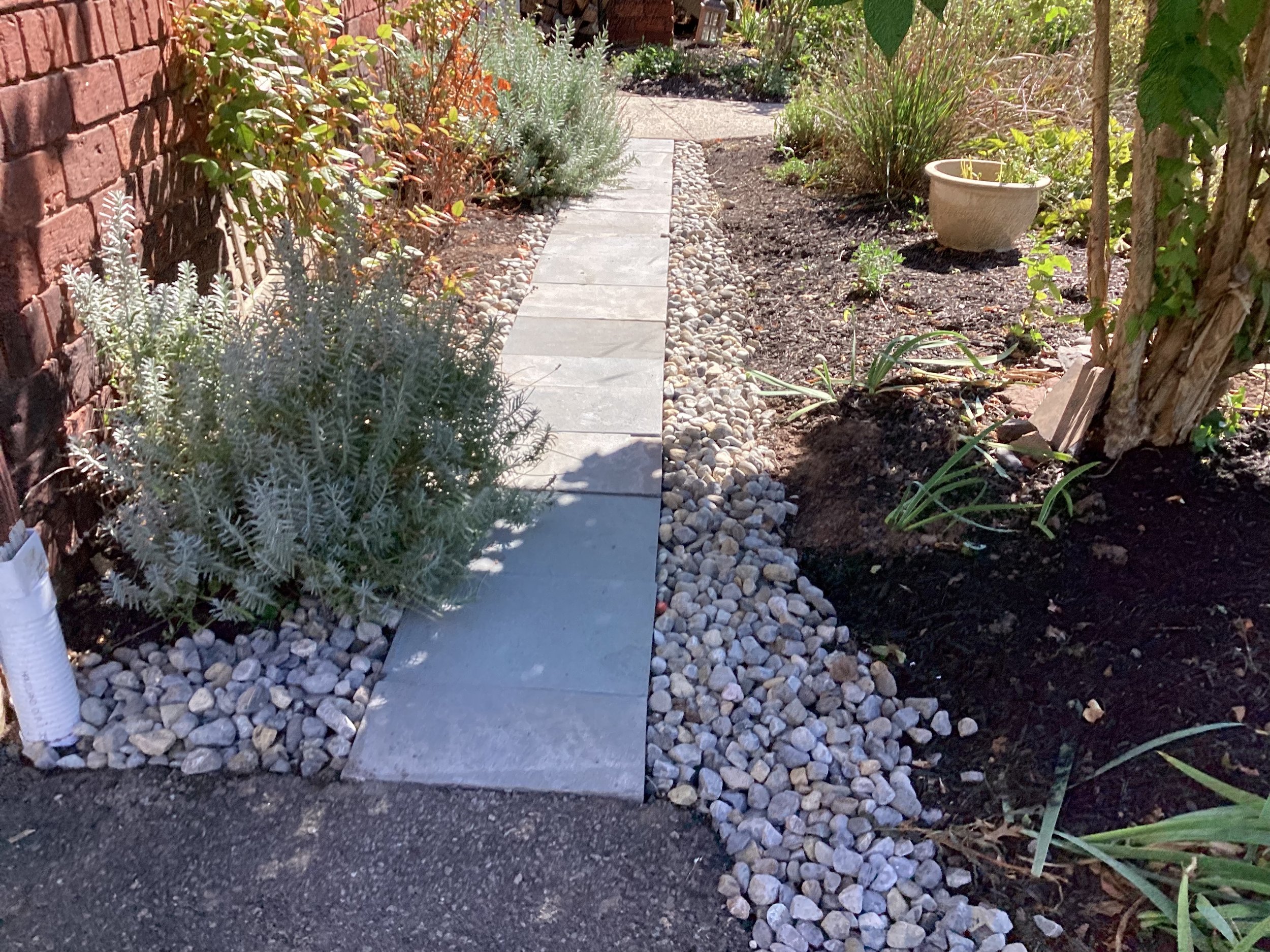 A garden pathway made with light gray paving stones, bordered by white decorative pebbles, with plants on both sides and a brick wall on the left.