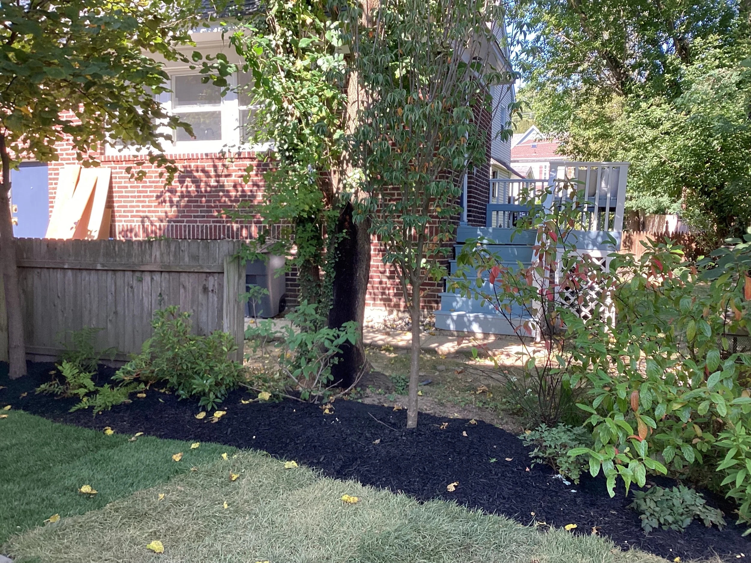 Backyard garden with trees, shrubs, a wooden fence, a brick house with a staircase and deck, and a grassy lawn.