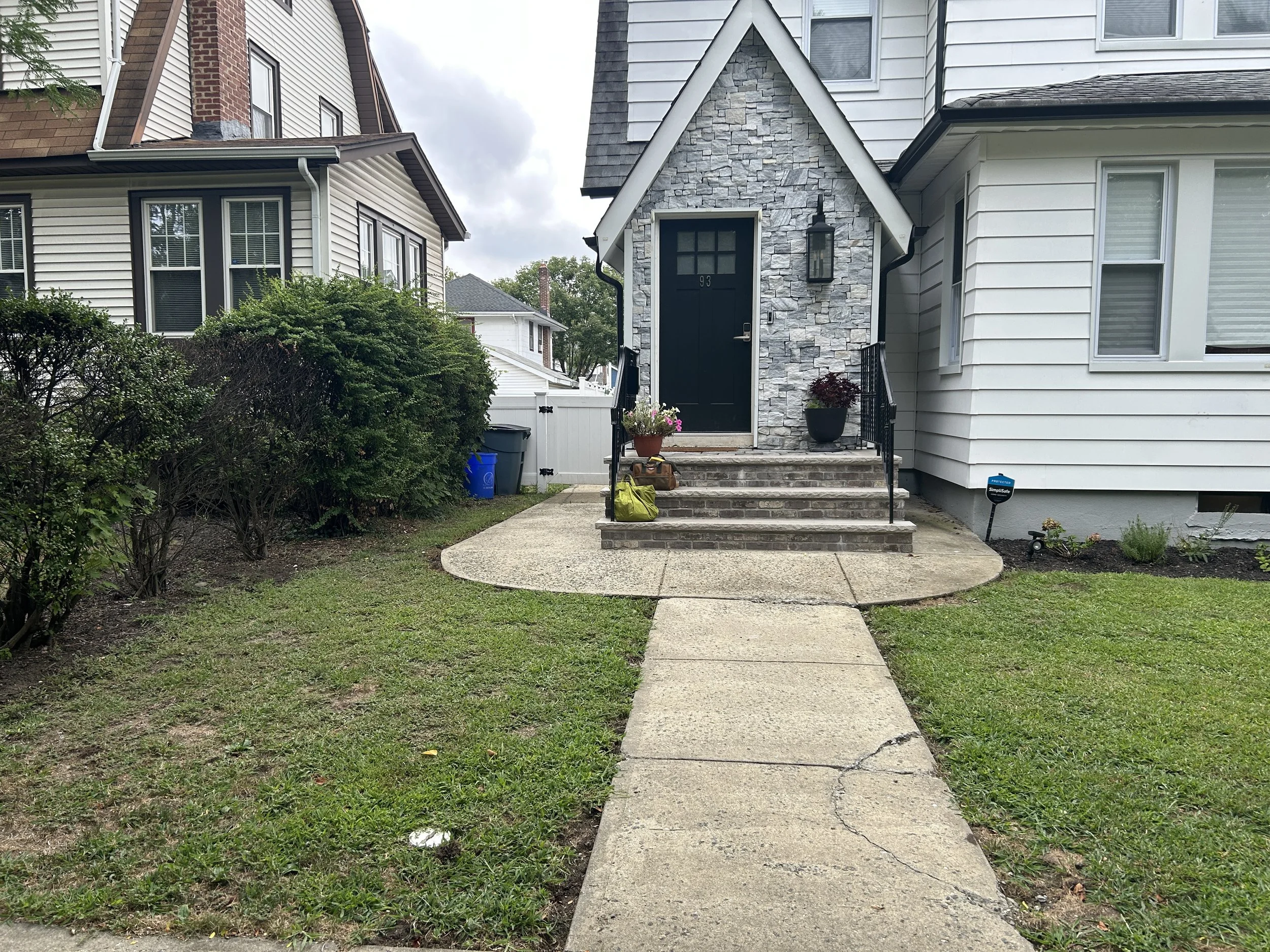 Front view of a house with a paved walkway leading to the stairs at the entrance, with green grass and bushes on either side, and potted plants on the porch.
