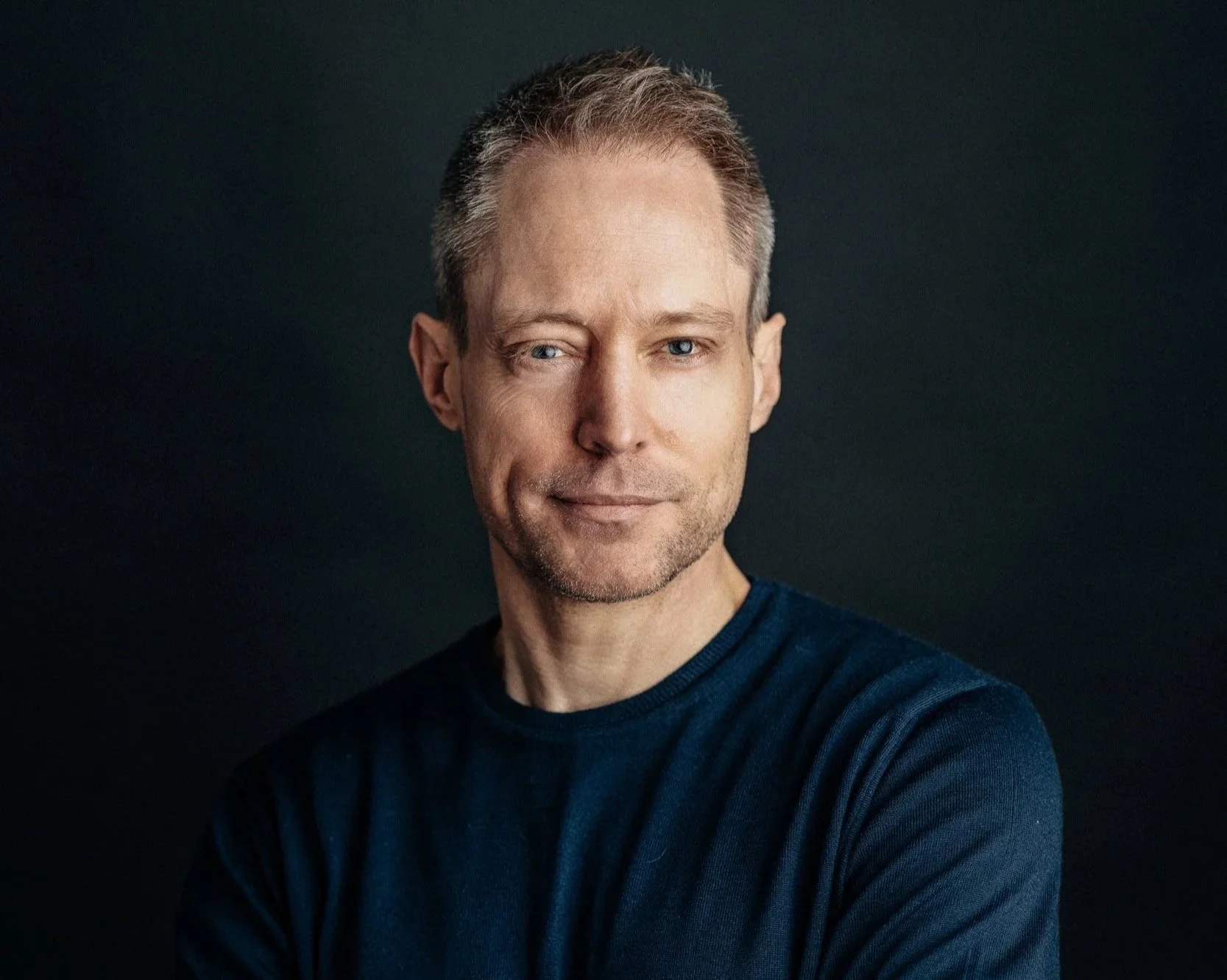 A man with short light brown hair, light skin, and blue eyes, wearing a navy blue shirt, smiling slightly, against a dark background.