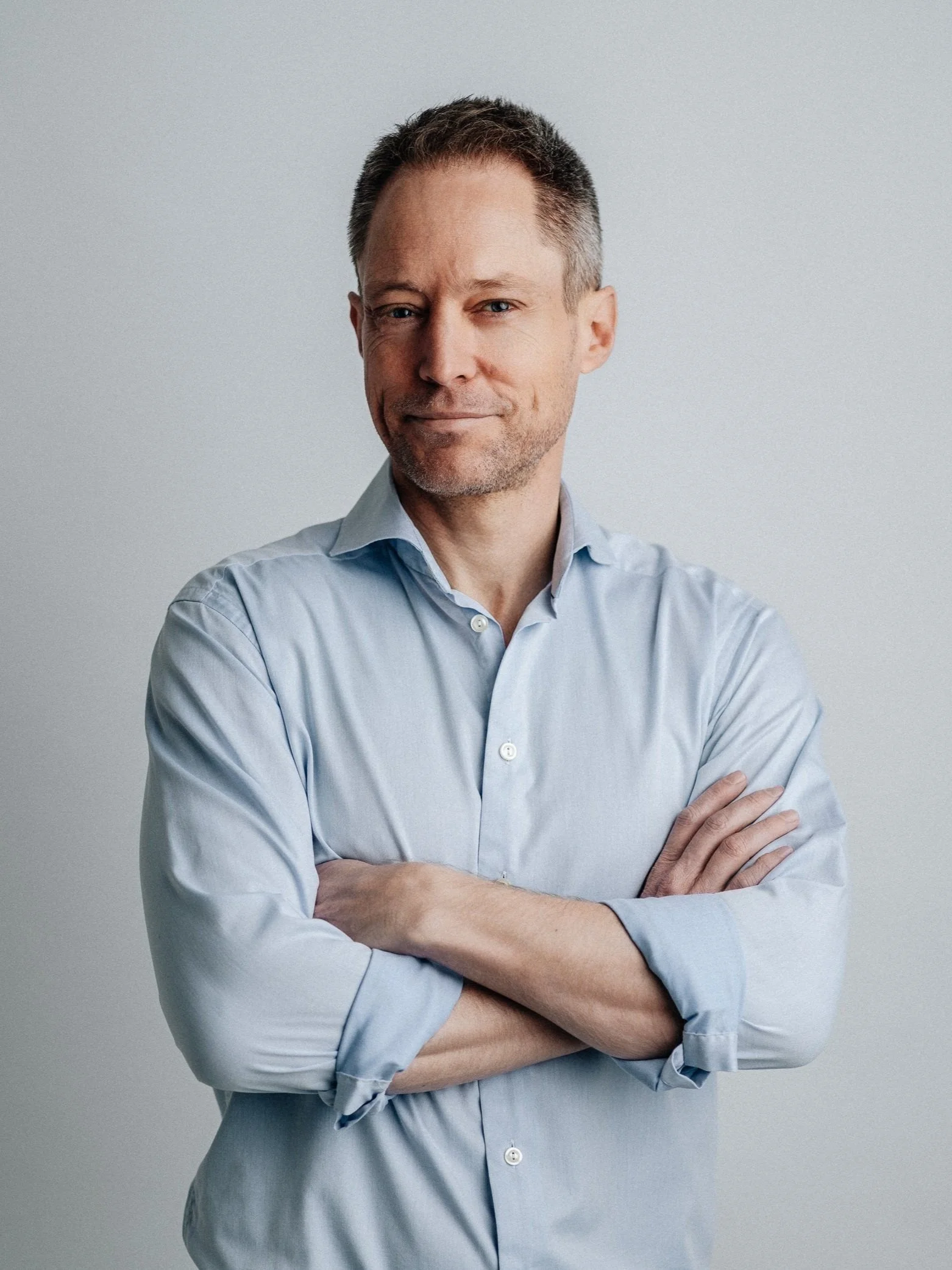 A confident man with short brown hair and slight facial stubble, wearing a light blue button-up shirt with rolled sleeves, standing against a plain light gray background.