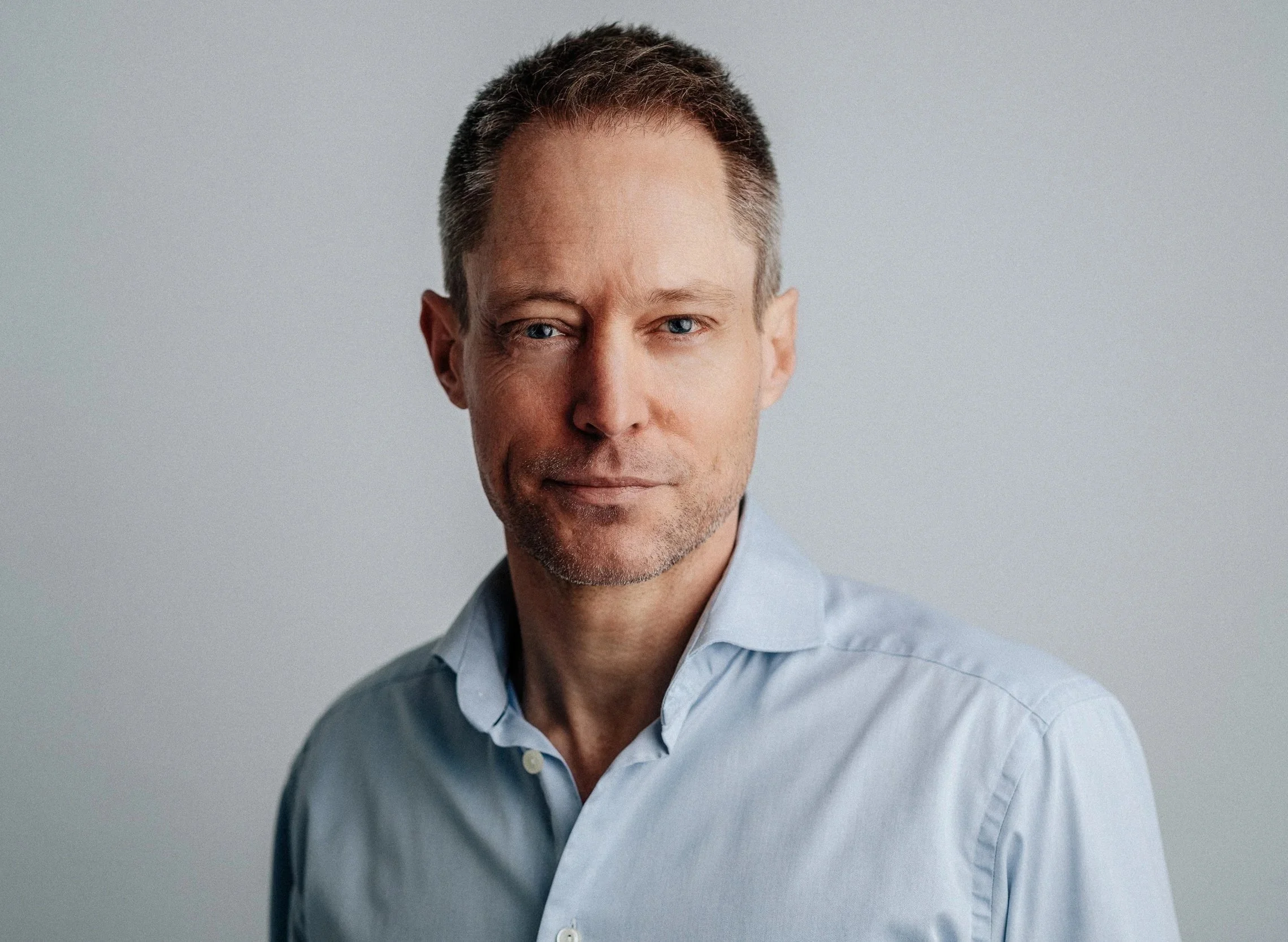 A middle-aged man with short, dark hair, blue eyes, and a slight beard, wearing a light blue button-up shirt, looking at the camera against a neutral background.