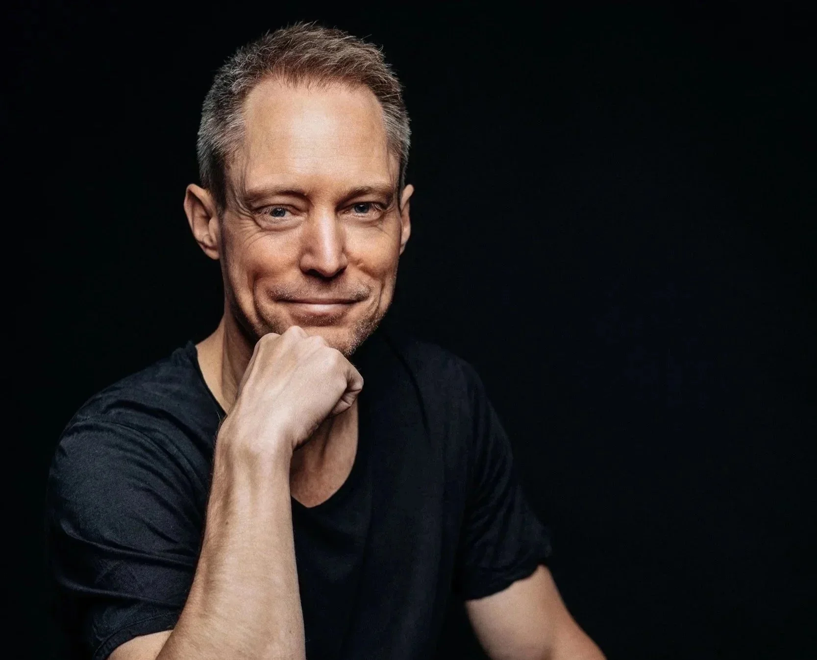 A middle-aged man with short gray hair, wearing a black t-shirt, smiling confidently with his hand resting on his chin against a black background.
