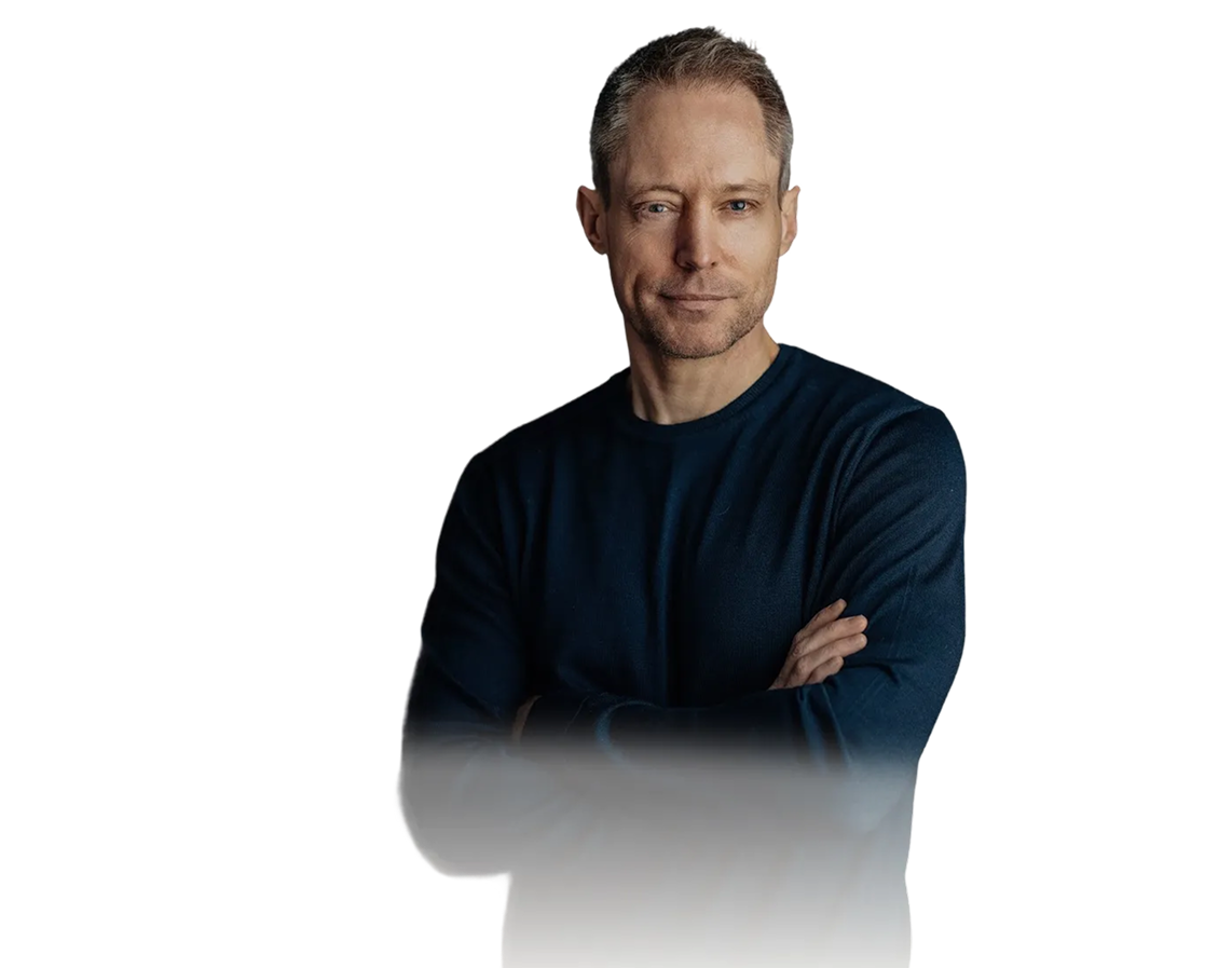 A man with short hair and a slight smile stands with arms crossed, wearing a dark long-sleeve shirt against a plain background.