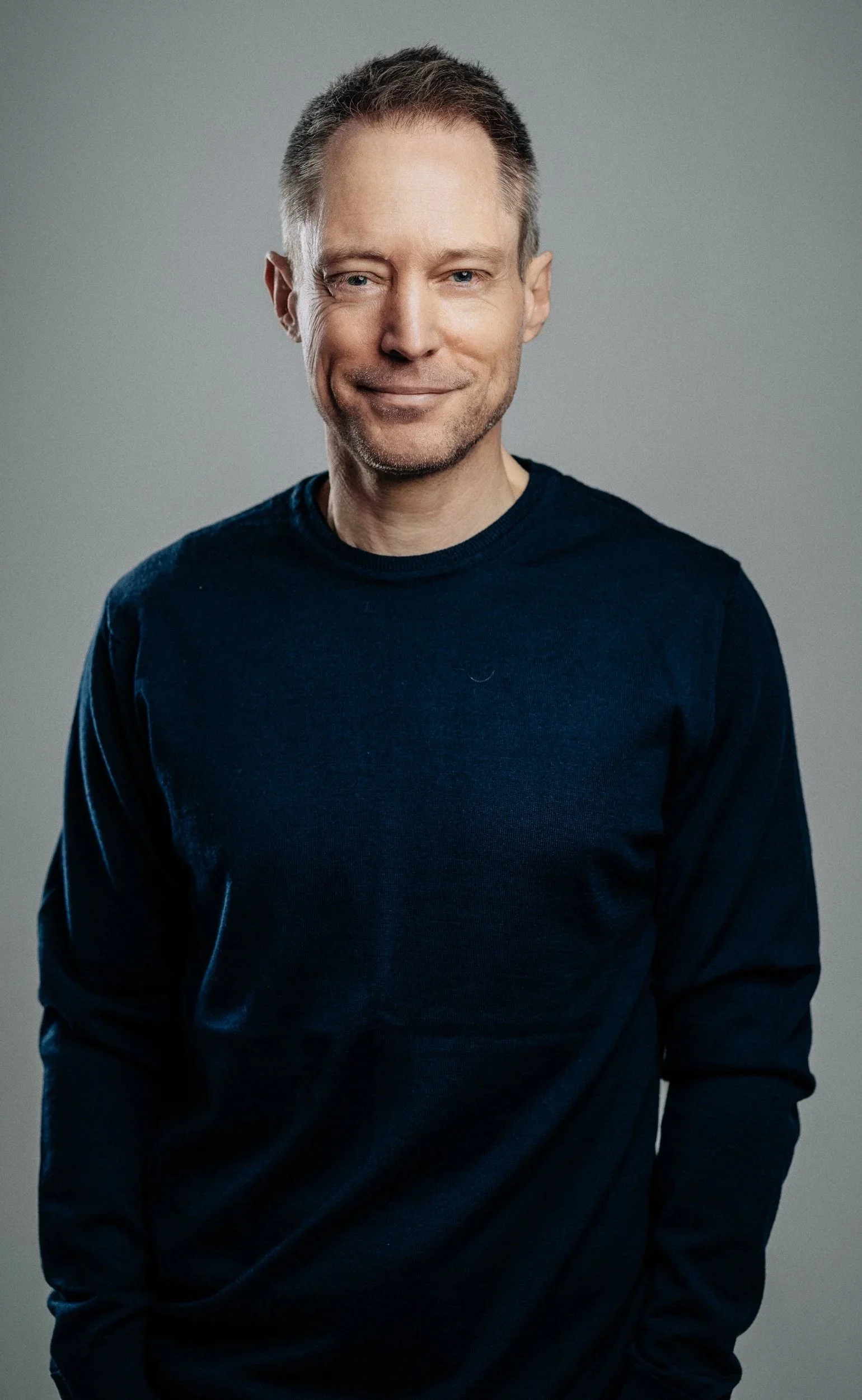 Portrait of a smiling middle-aged man with short brown hair, wearing a dark blue long-sleeve shirt, standing against a plain gray background.