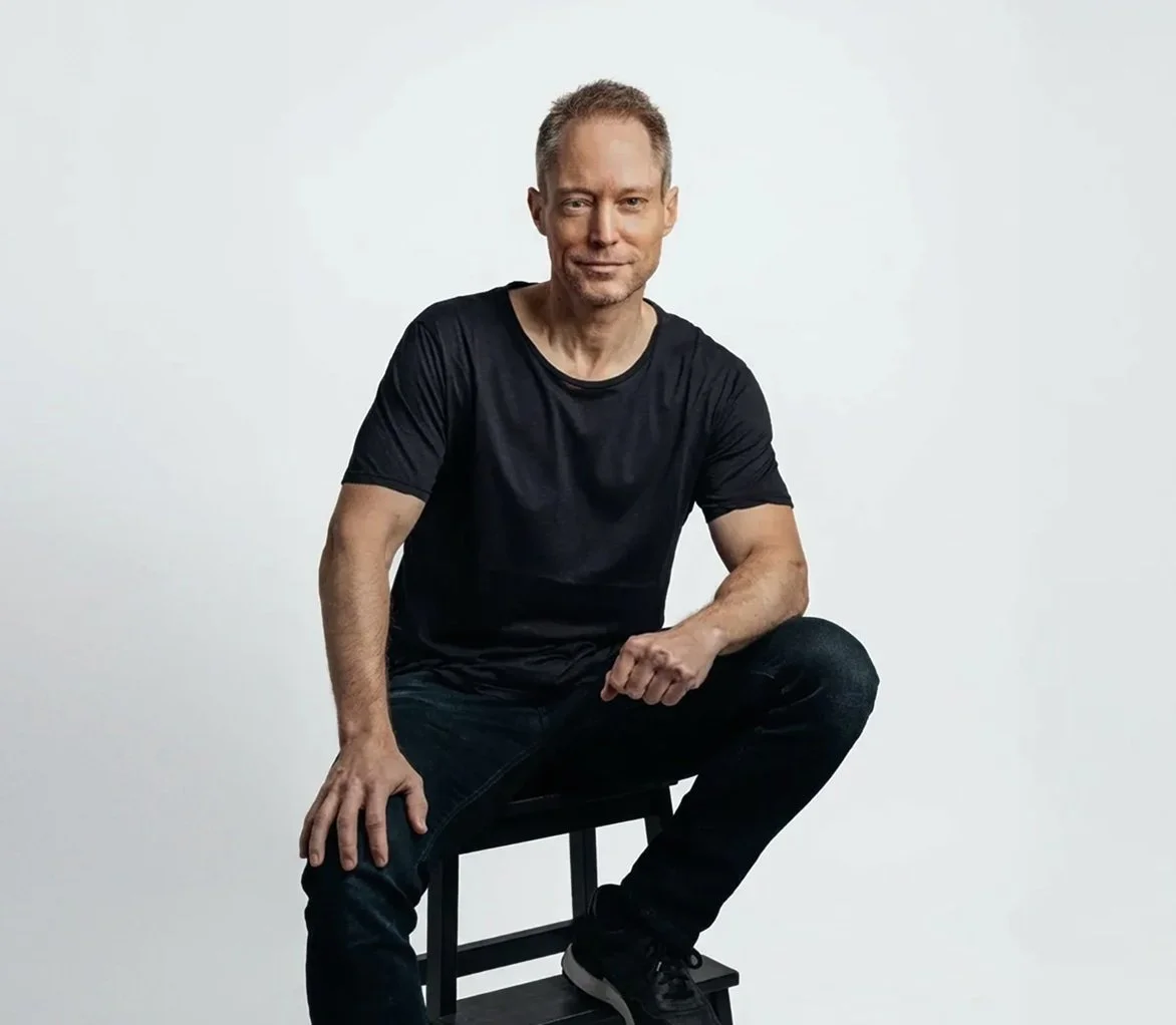A man in a black t-shirt and jeans sits on a stool against a plain white background, smiling at the camera.