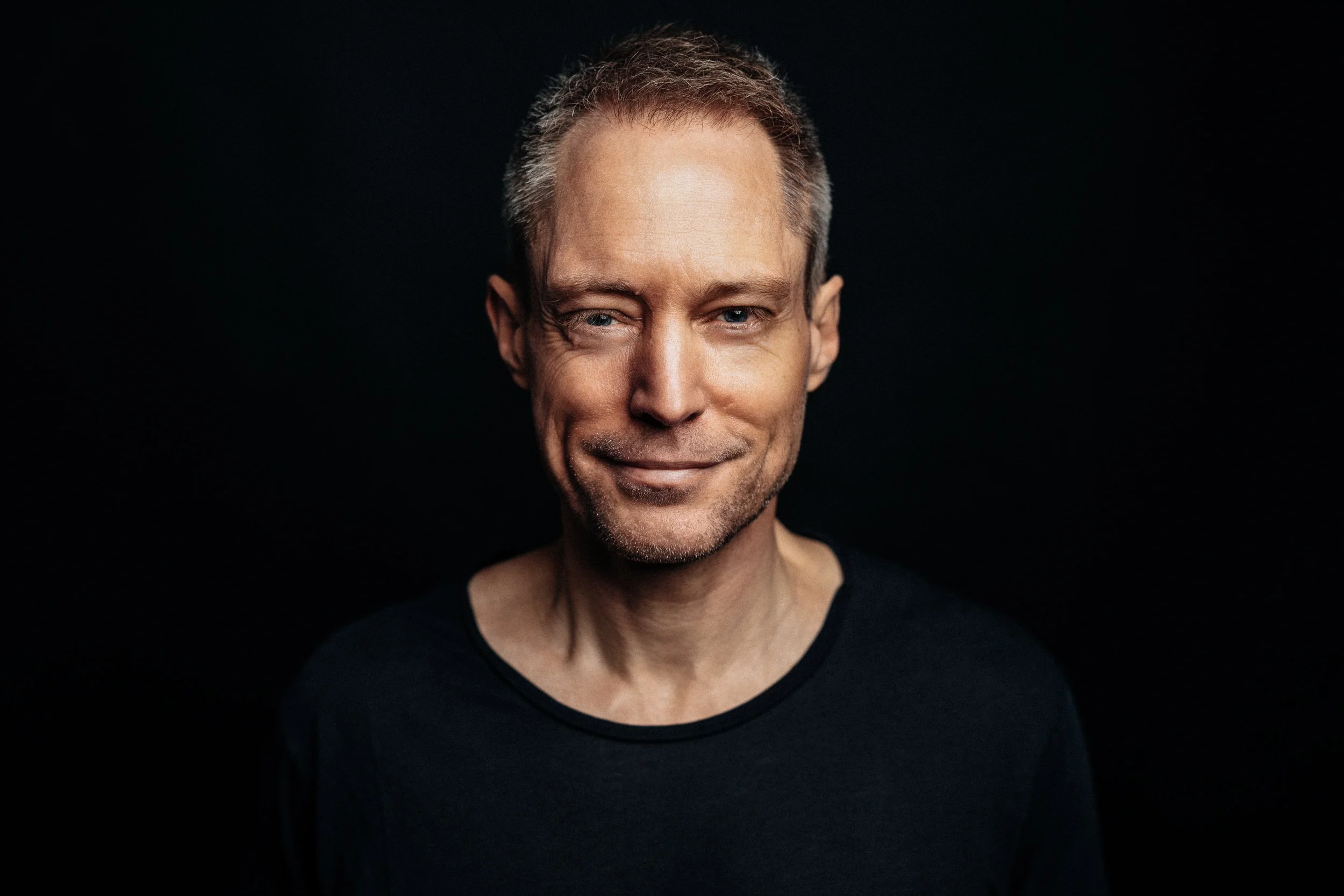 A middle-aged man with short, gray hair and blue eyes winks and smiles while wearing a black shirt against a dark background.