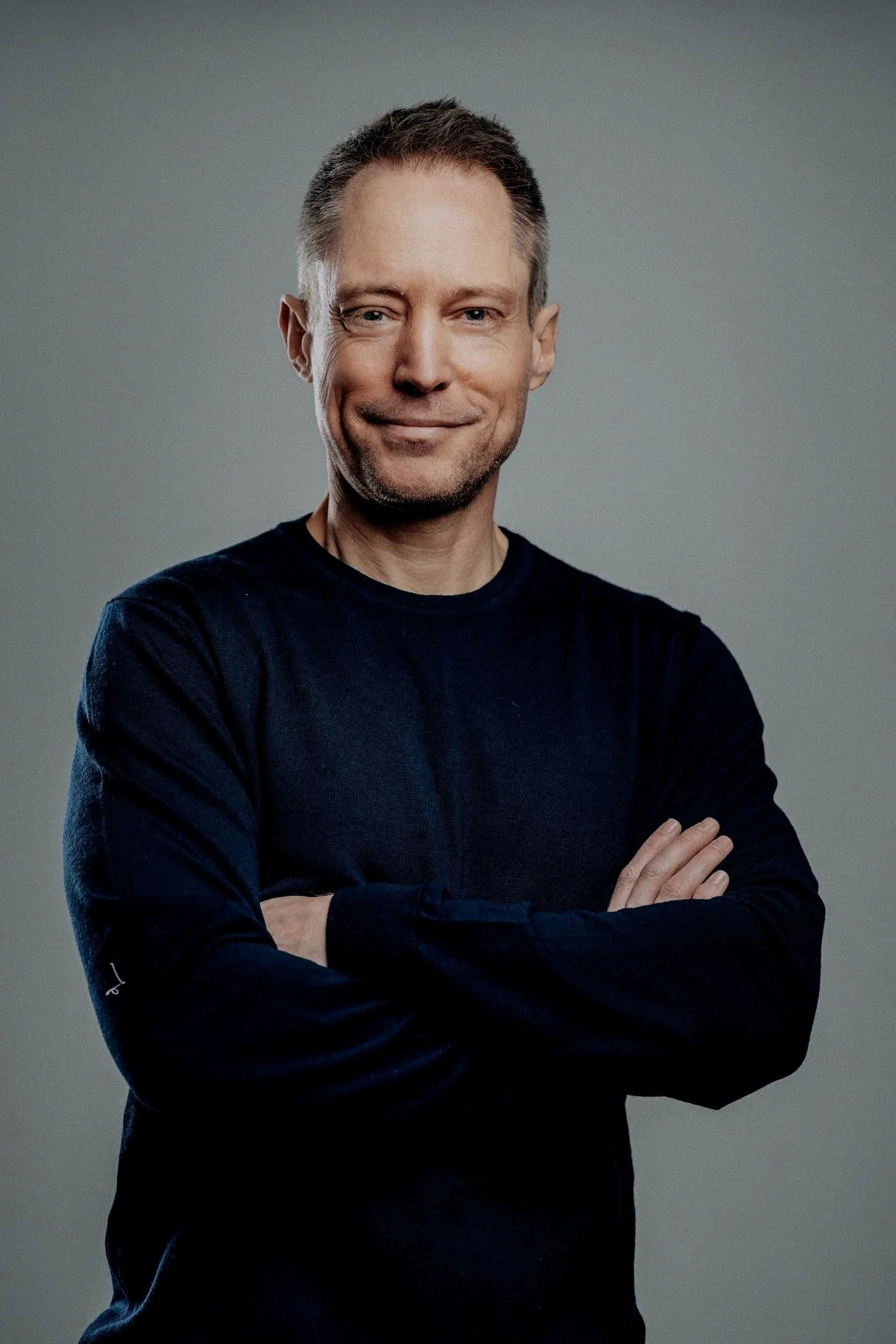 Portrait of a smiling middle-aged man with short hair, wearing a black long sleeve shirt, standing with arms crossed against a plain gray background.