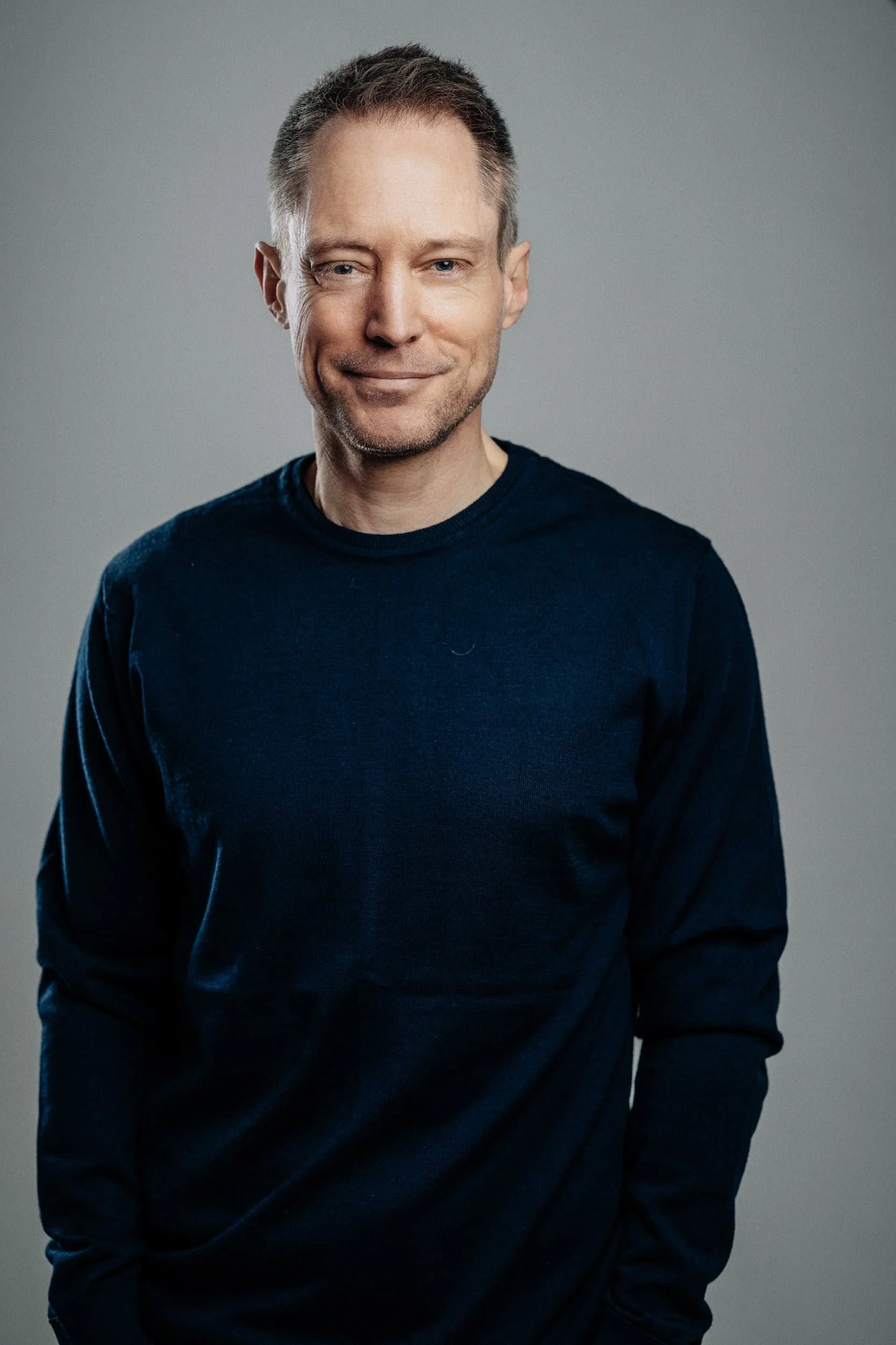 Portrait of a middle-aged man with short brown hair, wearing a dark blue long-sleeve shirt, smiling softly, against a plain gray background.