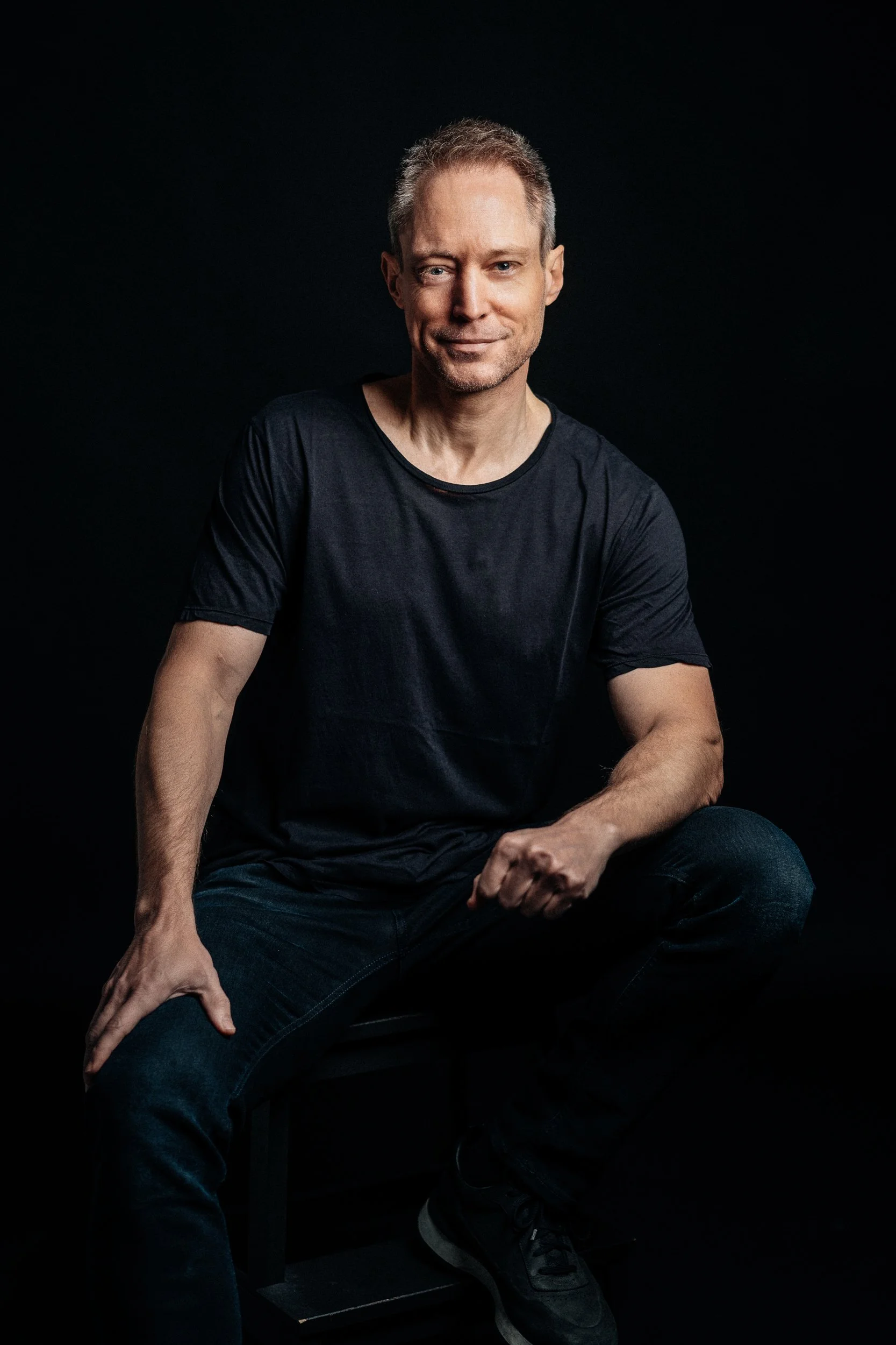 A man sitting on a black stool, wearing a black t-shirt and jeans, in front of a black background.