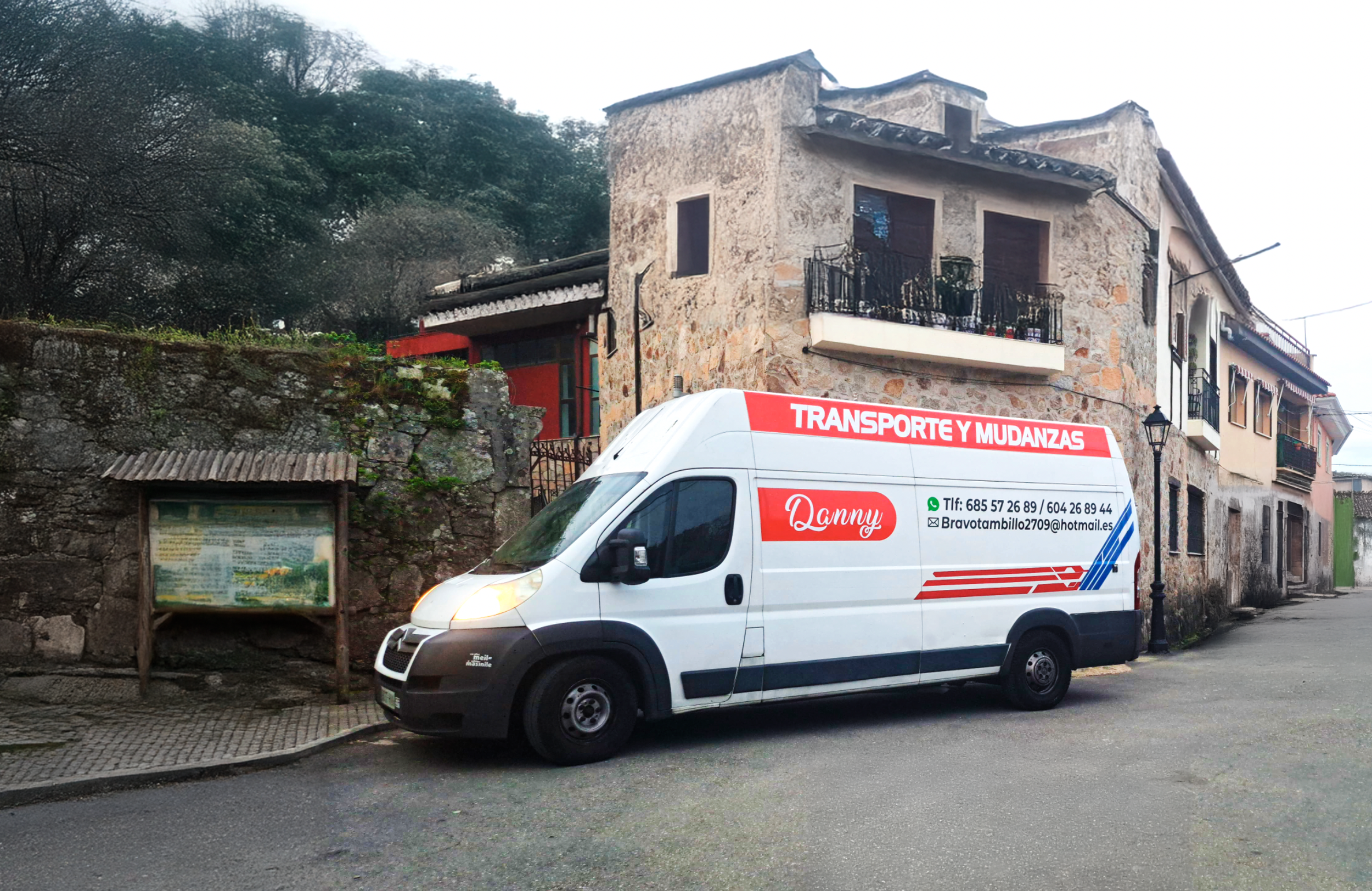 Camión de mudanzas blanco con letras rojas y negras, estacionado en una calle de un barrio con casas de piedra y paredes envejecidas, árboles en el fondo y una cartelera en la acera.