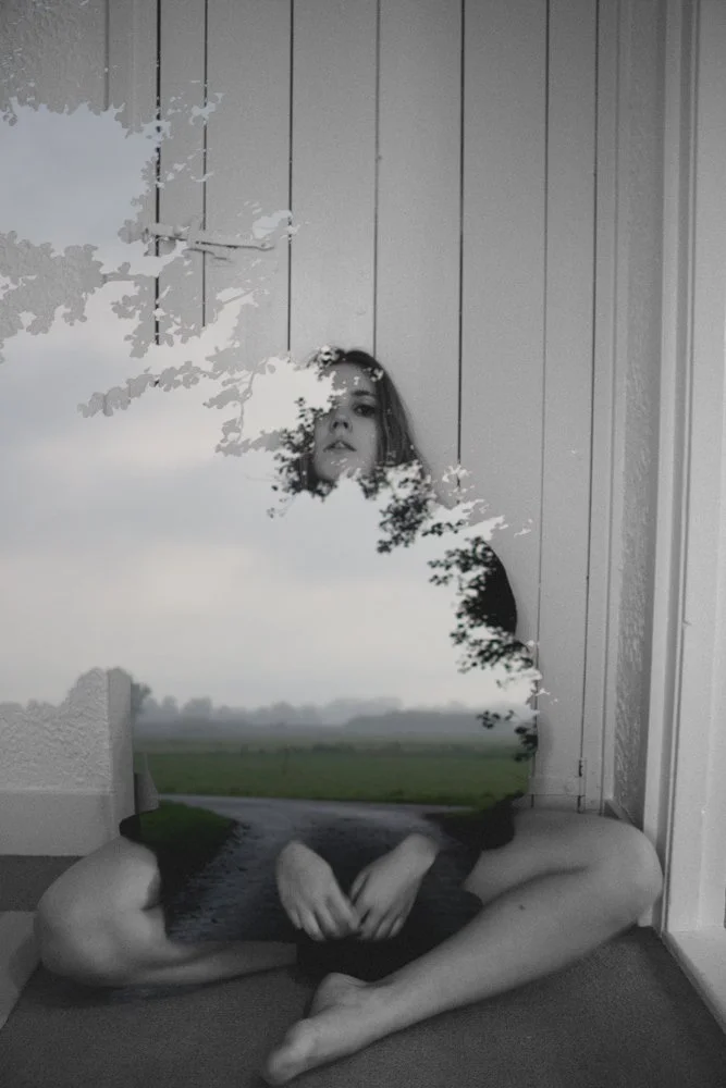 Een jonge vrouw zittend op de vloer in een hoek, met een spiegel die een landschap en de vrouw reflecteert.