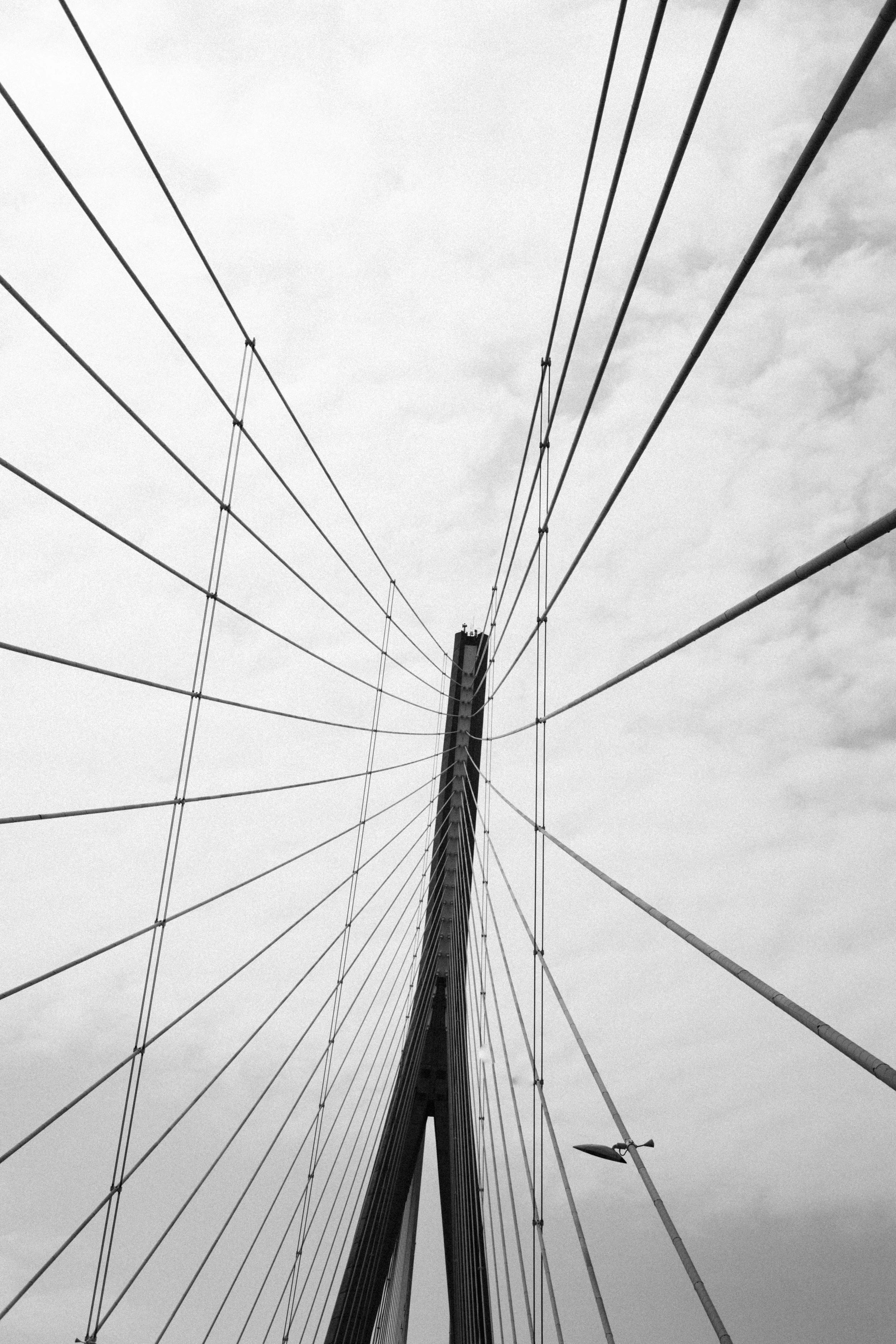 Close-up van een brug met draden die vanuit het midden naar de randen lopen, gezien vanaf onderen tegen een bewolkte hemel.