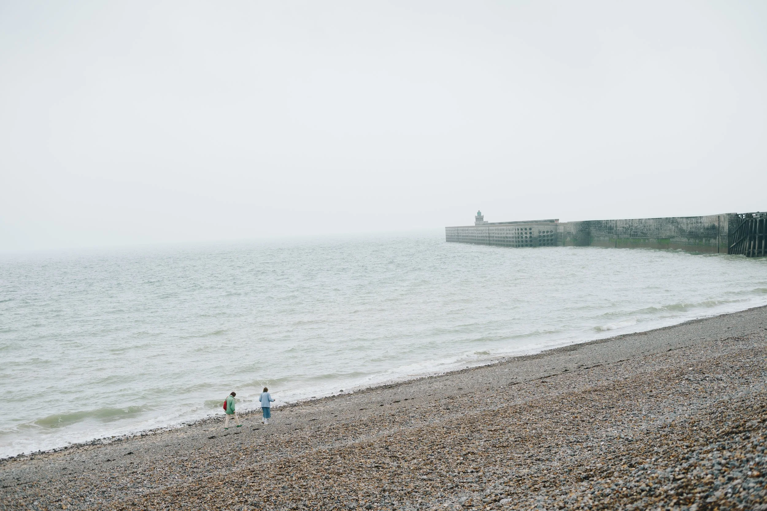 Twee mensen wandelen langs een kiezeldroge strand met een zee die onder een bewolkte, grijze hemel ligt, met een pier in de verte.