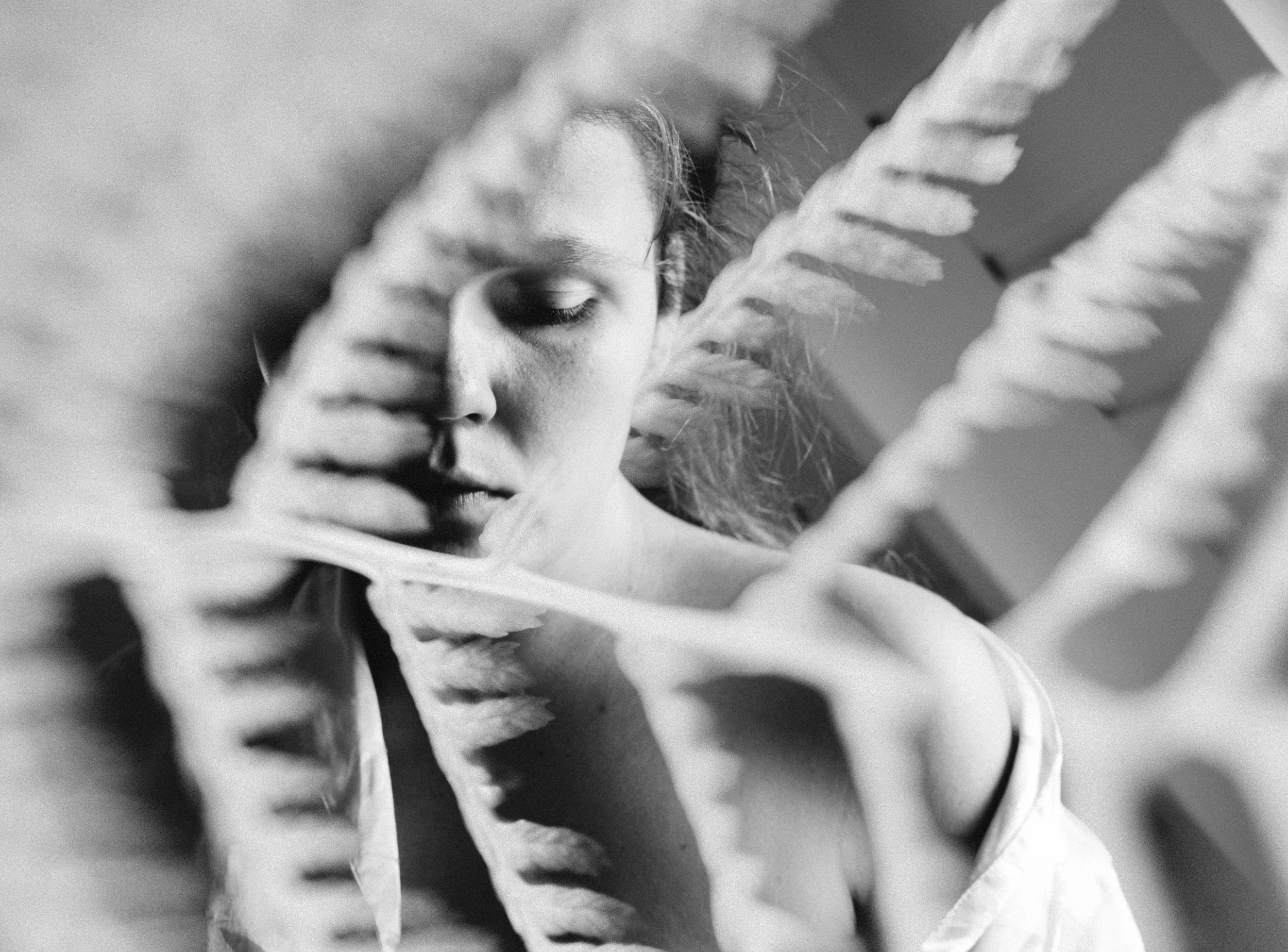 Zwart-wit foto van een vrouw die tussen grote bladeren van een plant kijkt, met haar gezicht gedeeltelijk bedekt door de bladeren.