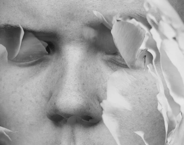Close-up zwart-wit van een sculptuur of beeld van een gezicht met bloembladeren waardoor de ogen gedeeltelijk verborgen zijn.