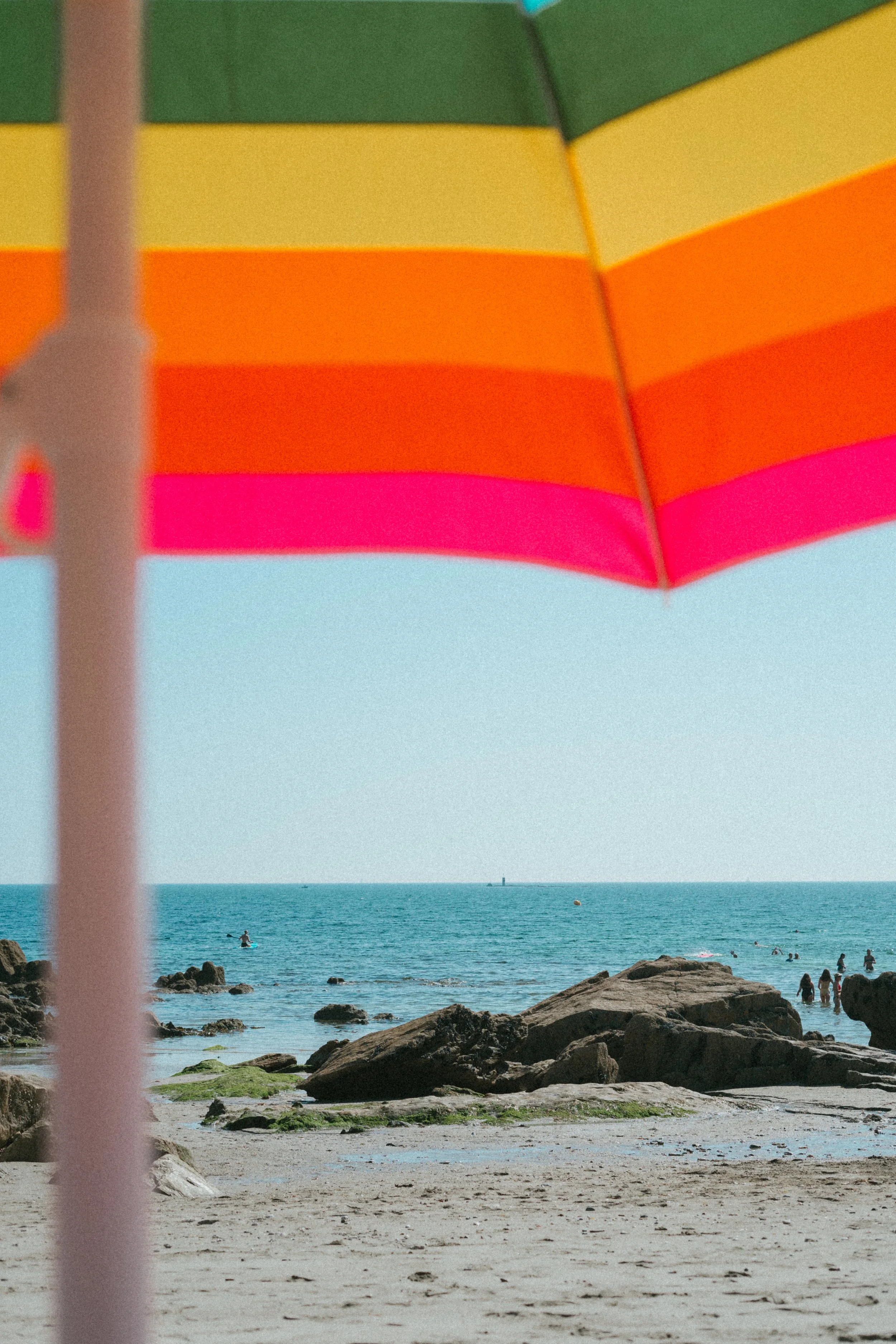 Strand met rotsen en mensen in de zee, een kleurrijke parasol op de voorgrond.