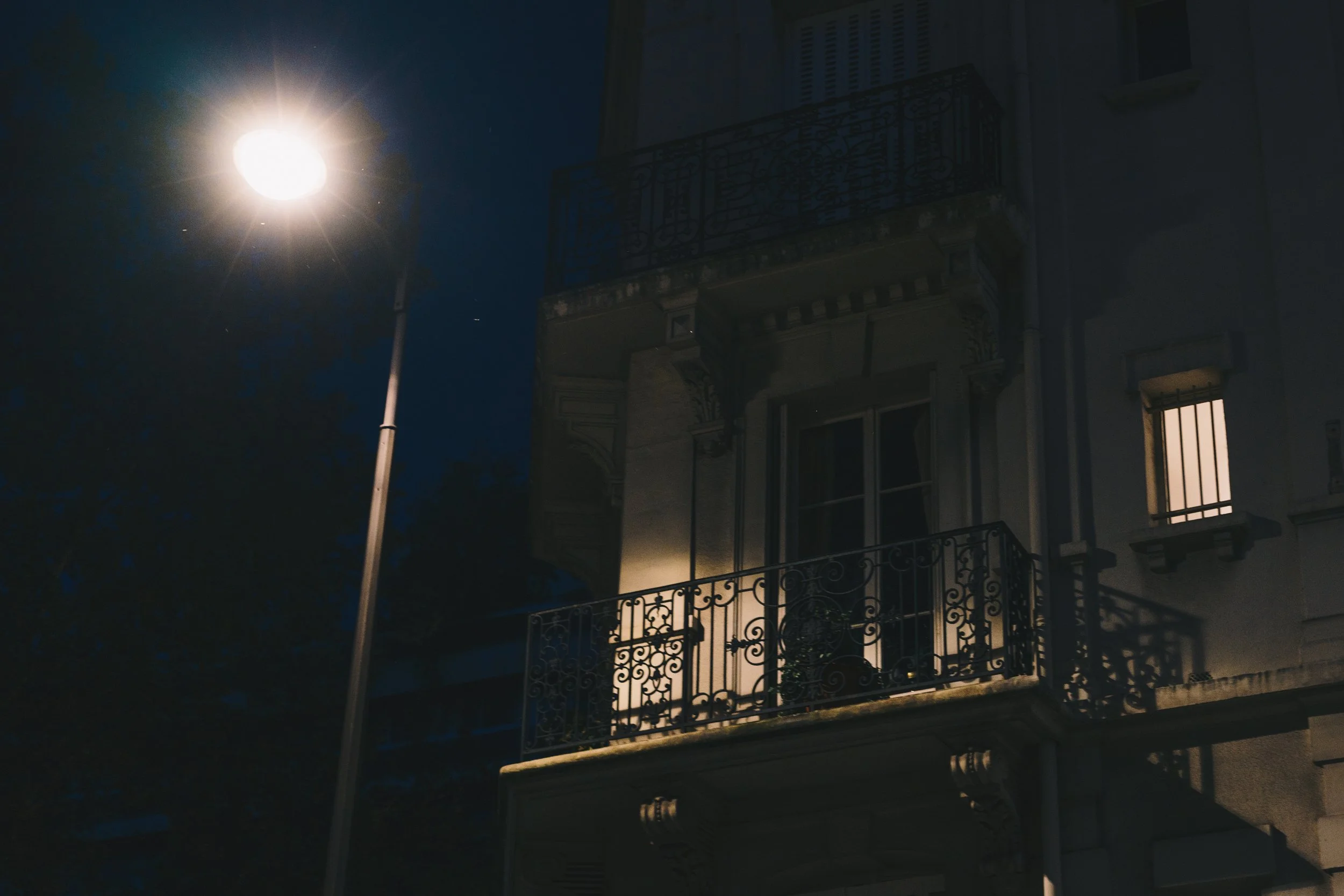 Een straatverlichting die 's nachts een balkon van een oud gebouw verlicht, met een raam met tralies en een licht binnen.