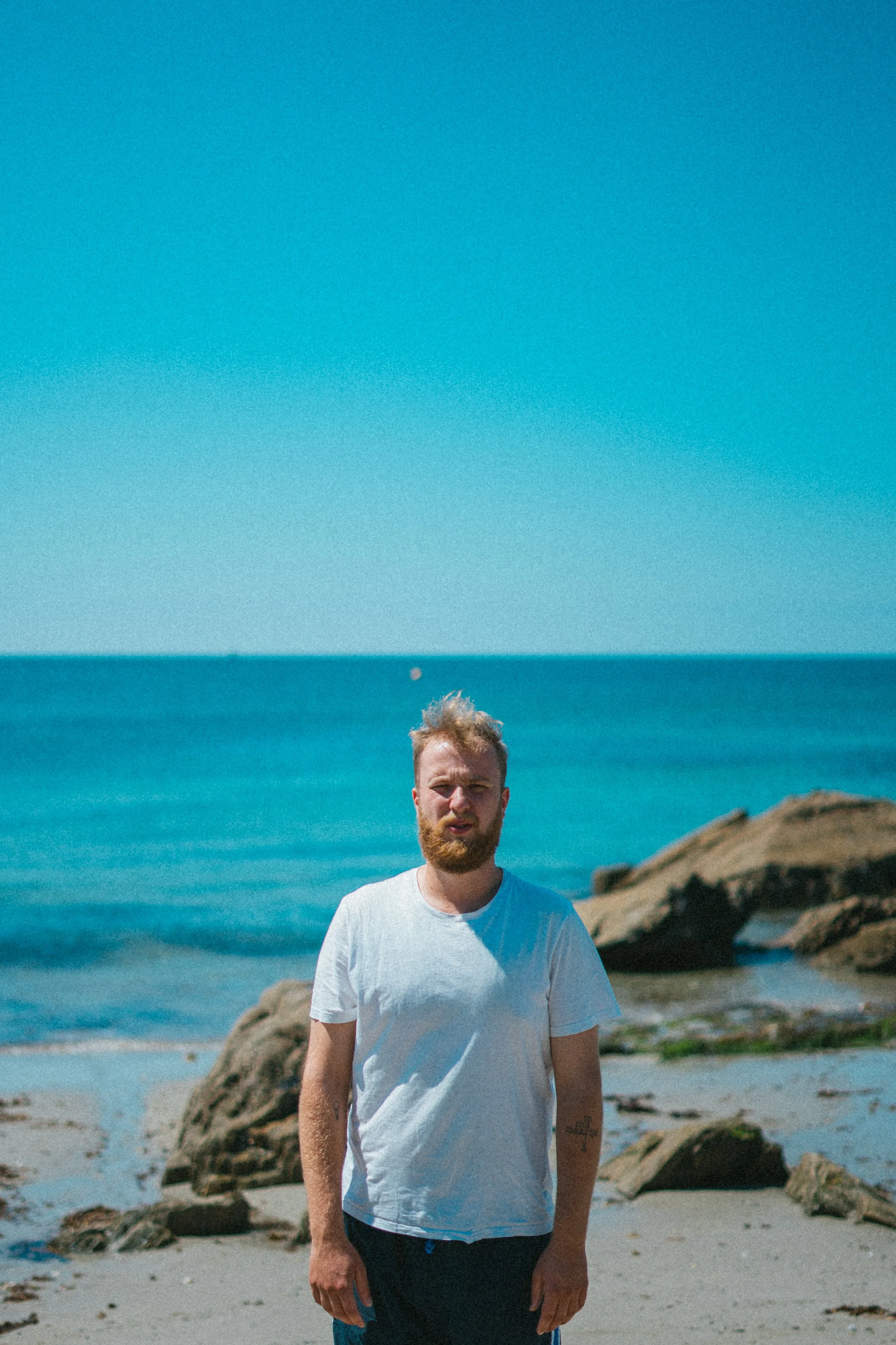 Man met een witte T-shirt op het strand met grote rotsen, de oceaan en een blauwe lucht op de achtergrond.