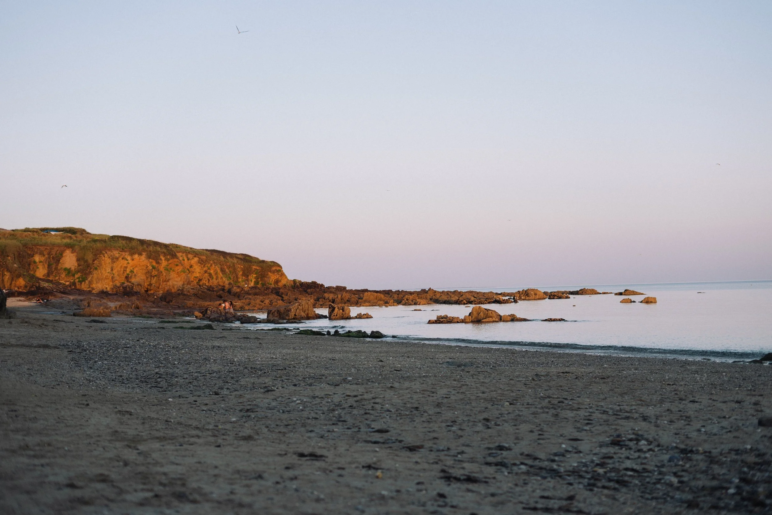 Foto van een rustiek strand met rotsen, een klif en een rustige zee onder een heldere hemel.