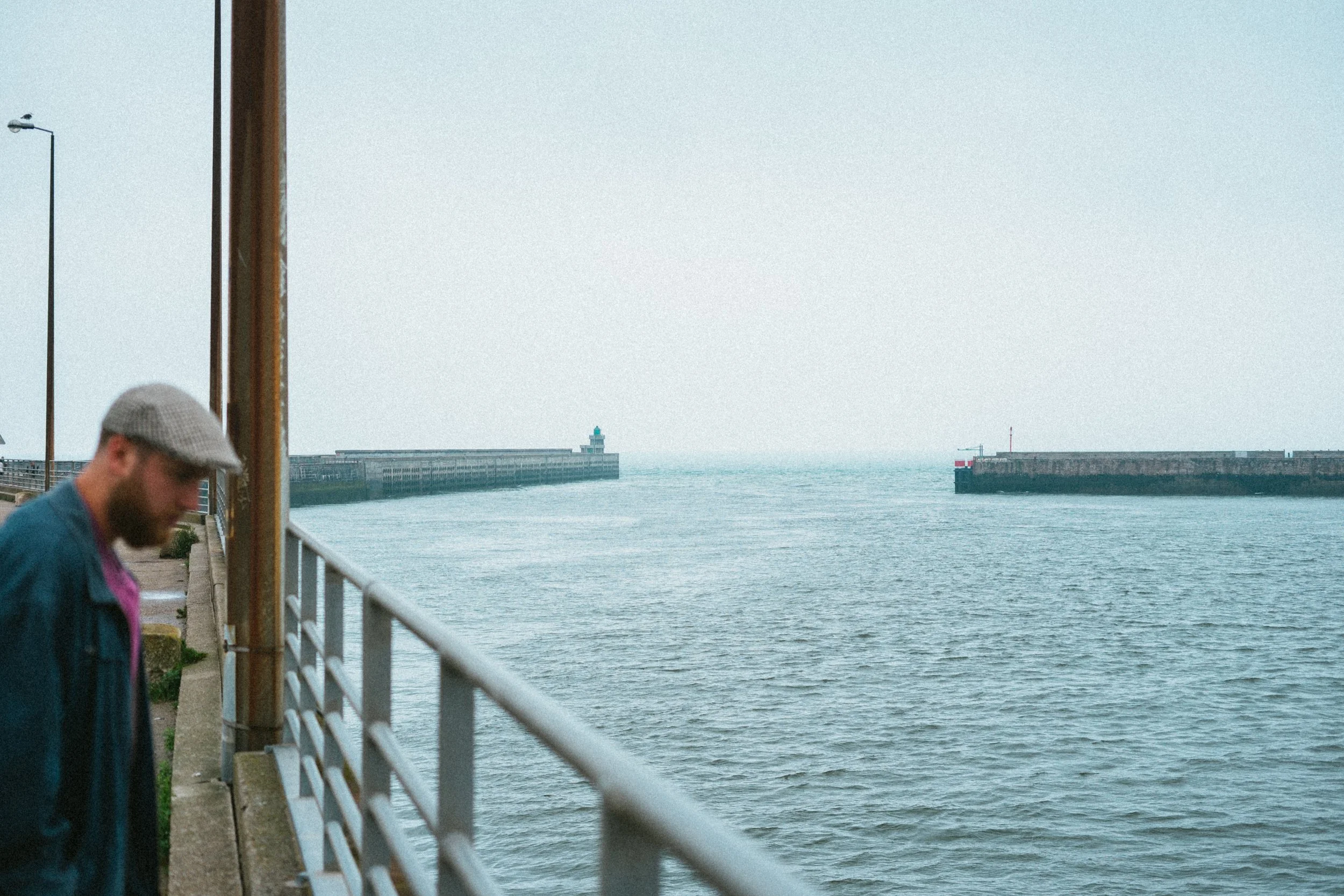Man in een pet en jas wandelt langs de kade bij de zee, met havenmuren en een vuurtoren op de achtergrond.