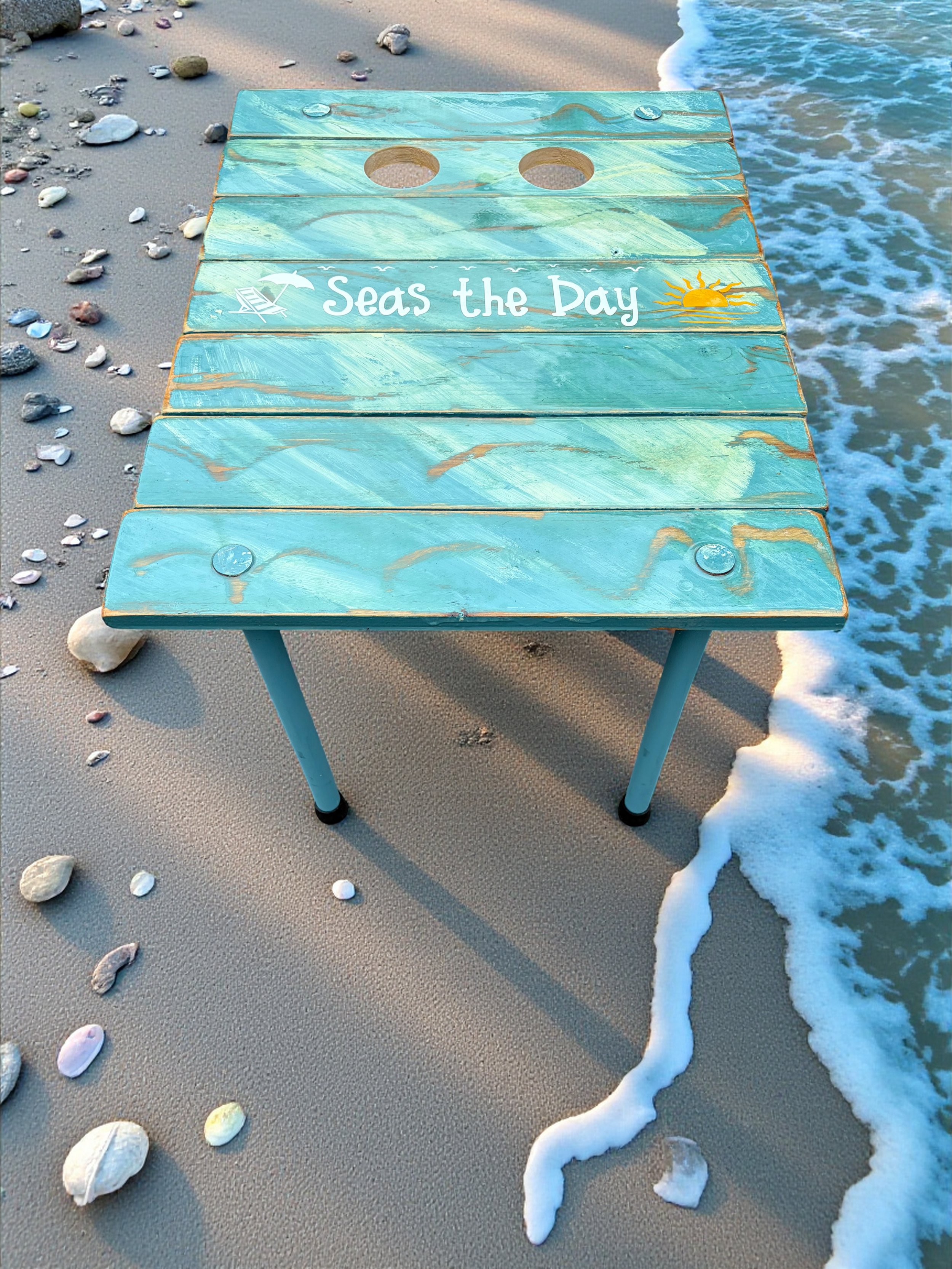 a turquoise beach table that rolls up and says seas the day on a sandy beach with cup holders