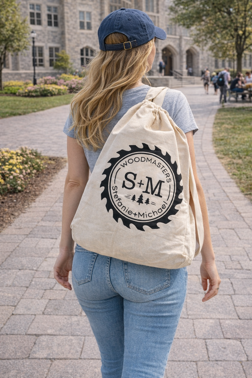 Woman walking on a college campus wearing jeans and a baseball cap, carrying a Woodmasters canvas drawstring bag with logo