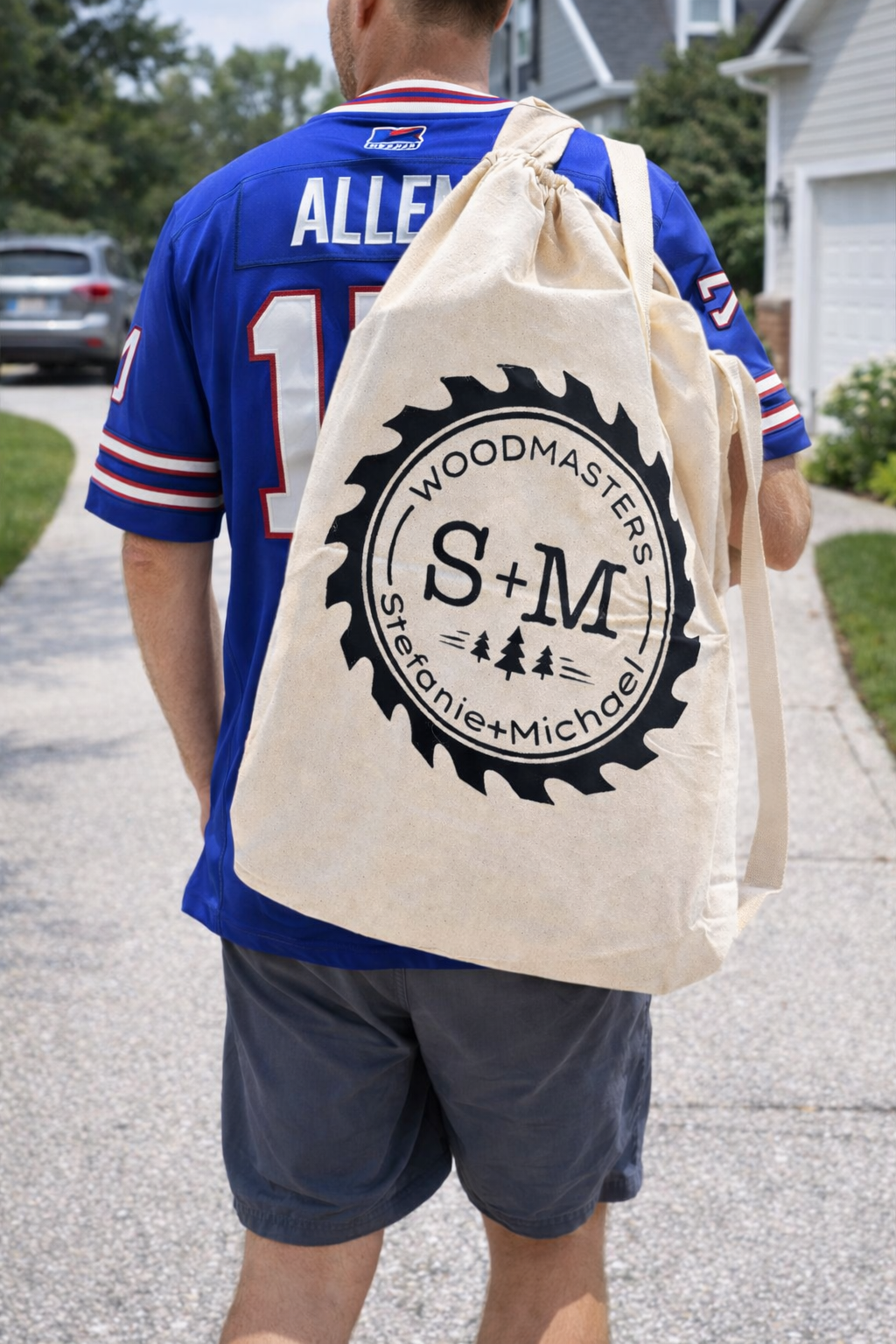 Man wearing a blue sports jersey with 'Allen' and '10' on the back, carrying a large beige tote bag with 'WOODMASTERS S+M Stephanie+Michael' and a gear logo with trees, sidewalks, a driveway, and houses in the background.