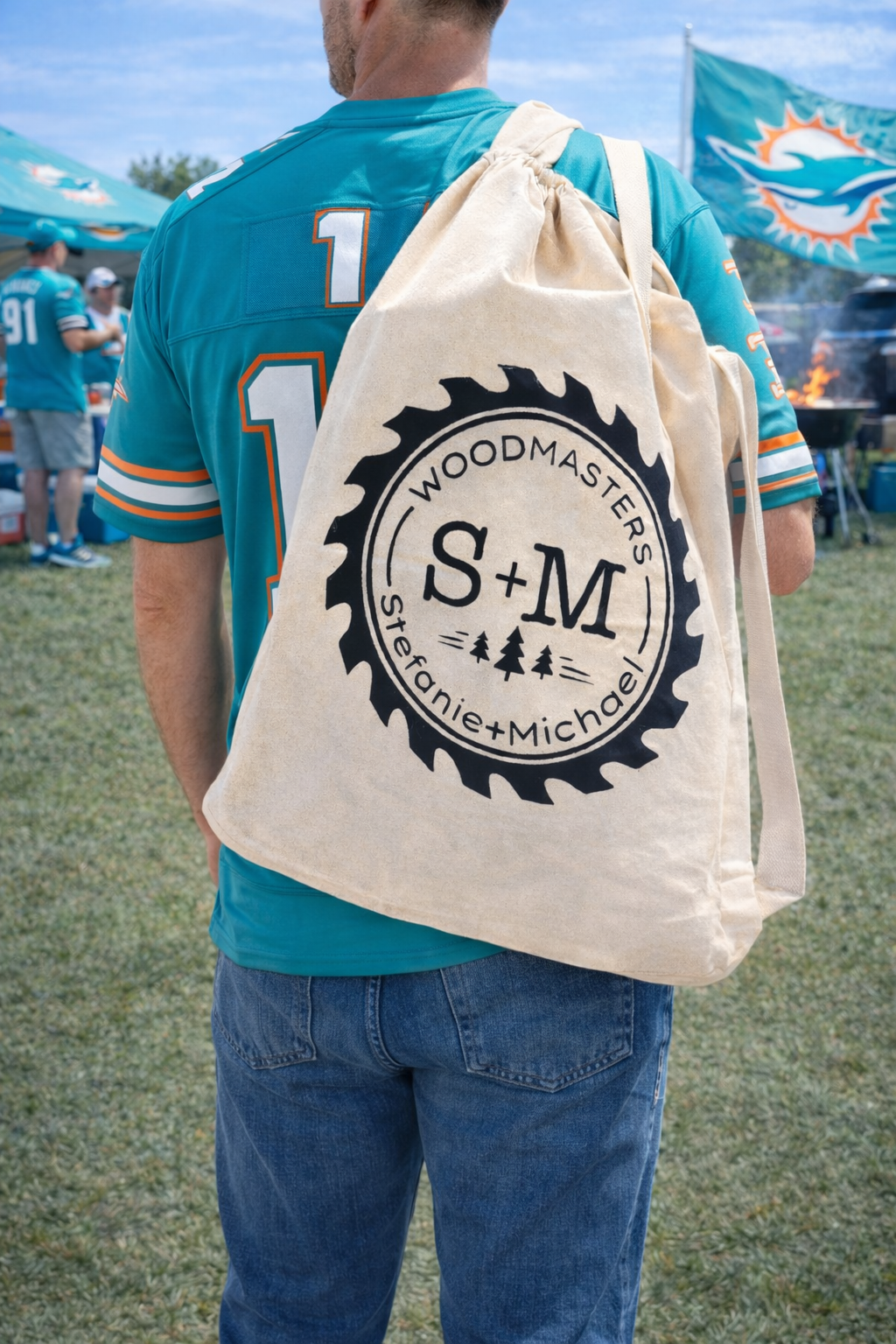 Man wearing Miami Dolphins jersey and jeans carrying S+M Woodmasters canvas table bag at a Dolphins tailgating scene