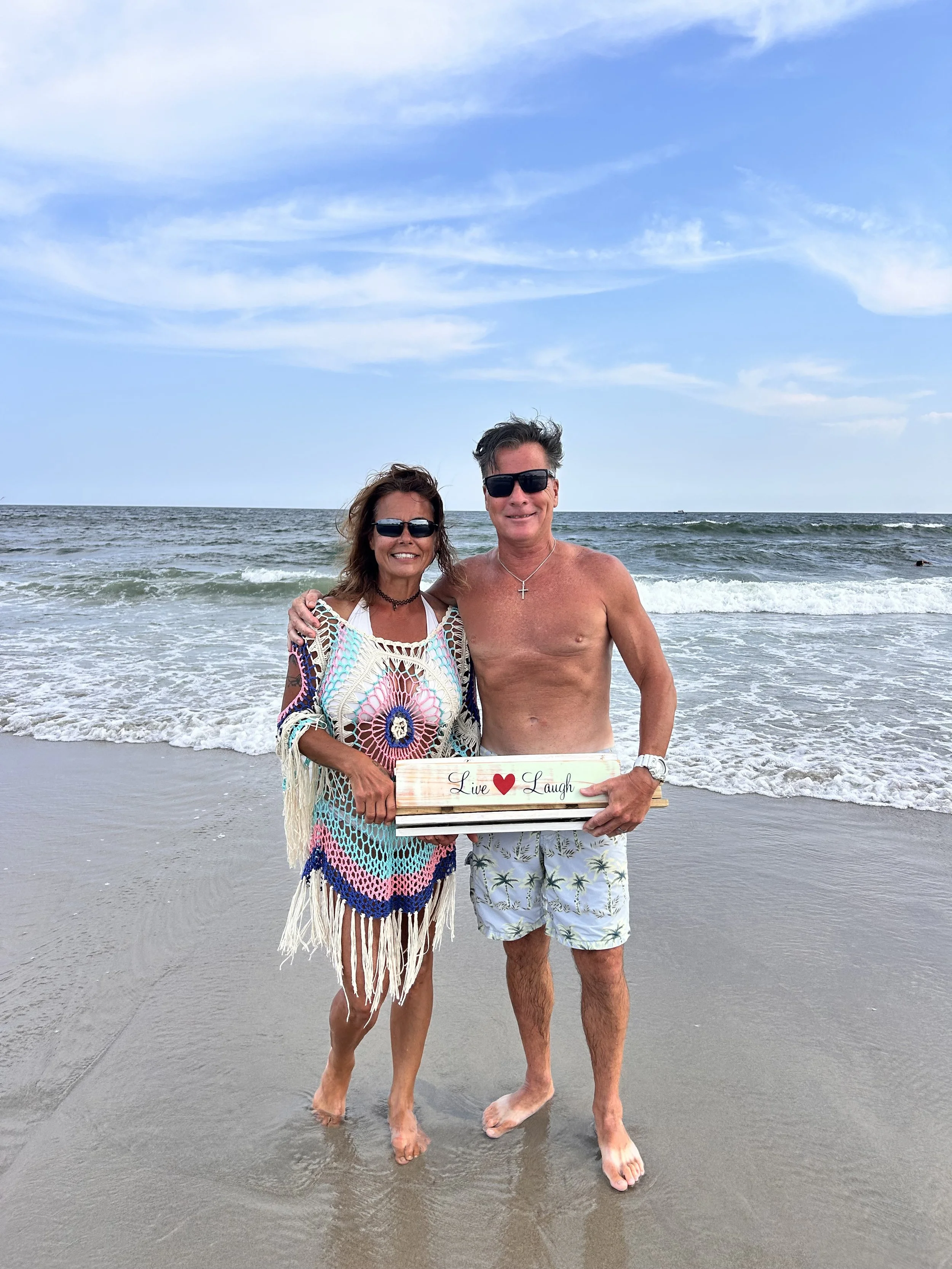 A smiling woman and man standing barefoot on the beach near the ocean, holding a decorative sign that says 'Live Love Laugh.' The woman is wearing a colorful, crocheted cover-up over a swimsuit, and the man is shirtless wearing swim trunks with a palm tree pattern. They are both wearing sunglasses, and the sky above is partly cloudy.