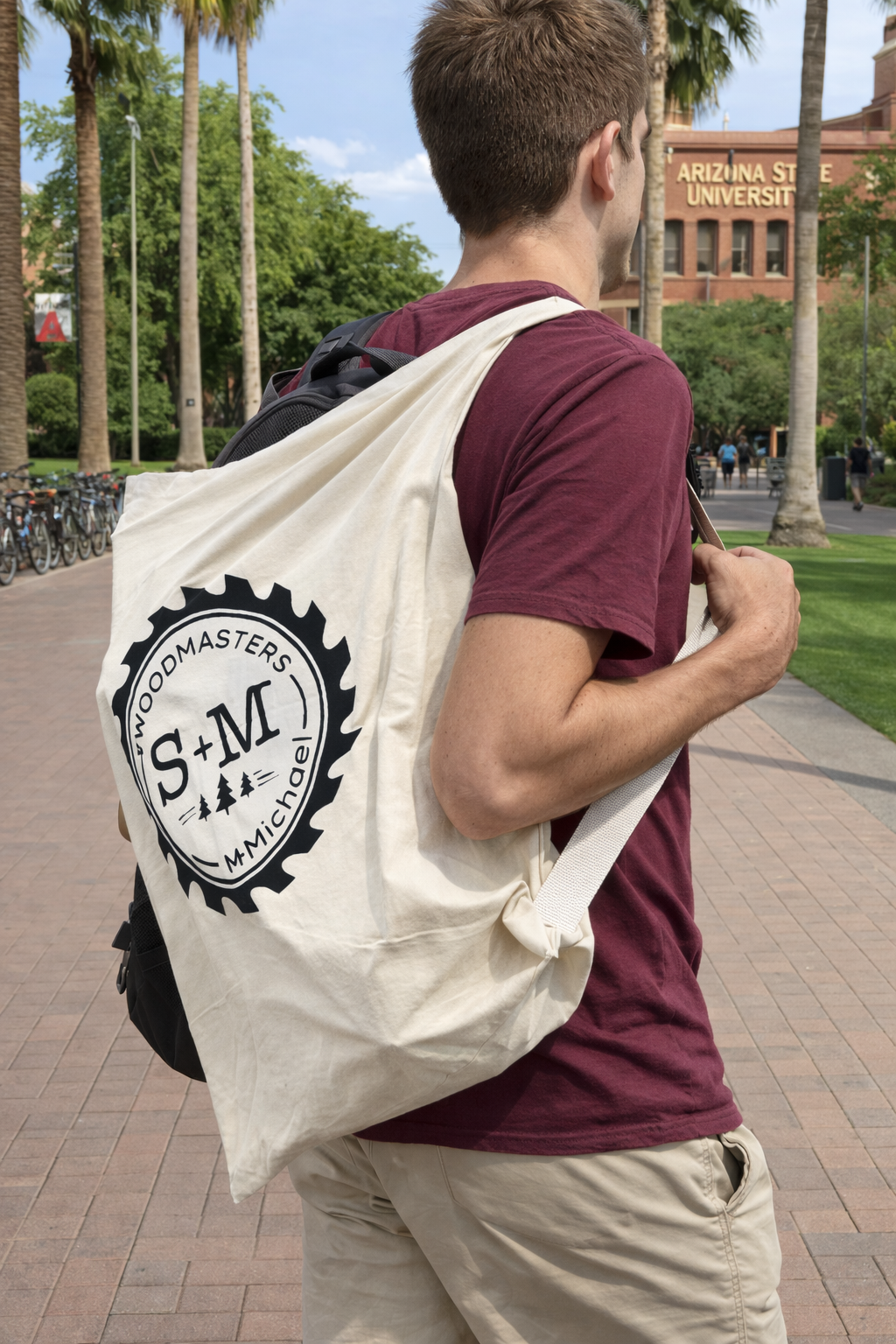 College student carrying S+M Woodmasters canvas tote bag while walking on an Arizona university campus with palm trees