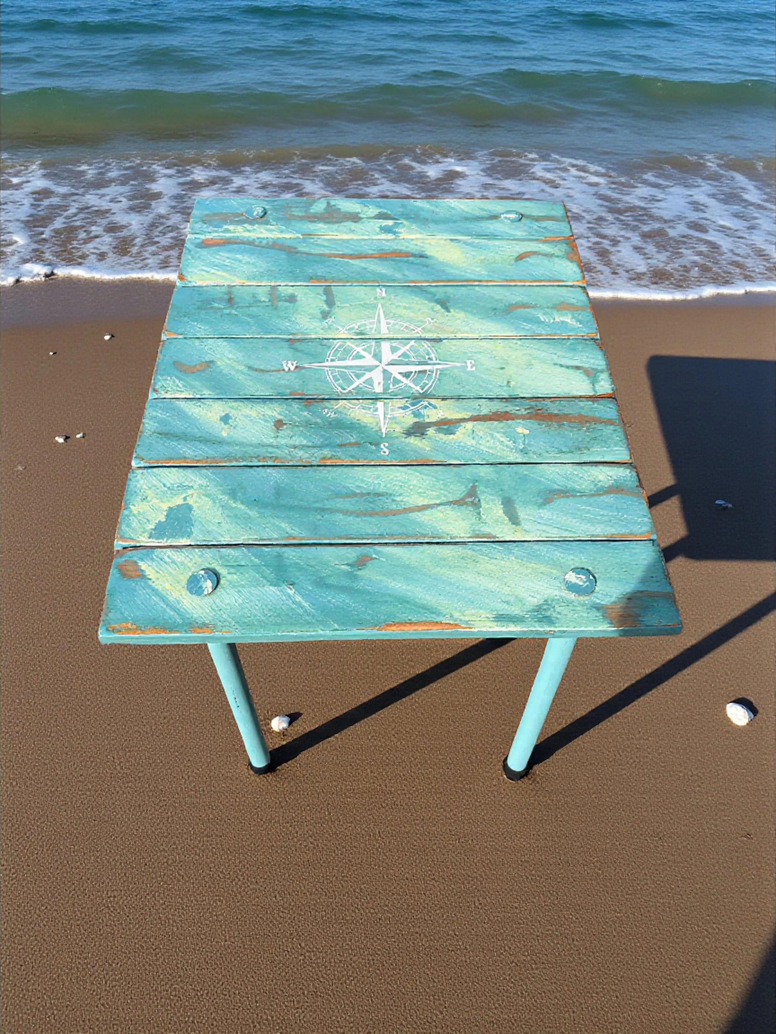 turquoise portable beach table with a white compass rose on the beach with shells and small waves