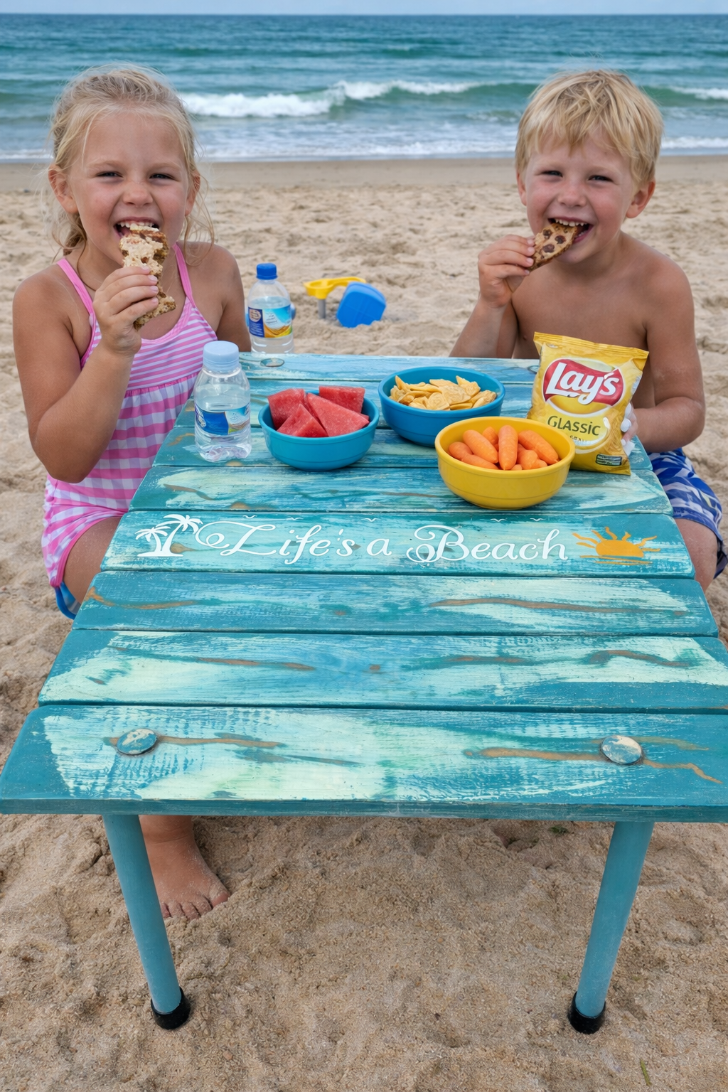 Two children, a girl in a pink striped swimsuit and a boy in blue shorts, sitting at a blue wooden table on the beach, eating cookies and chips, with bowls of watermelon, chips, and carrots, and water bottles. The table has the phrase "Life's a Beach