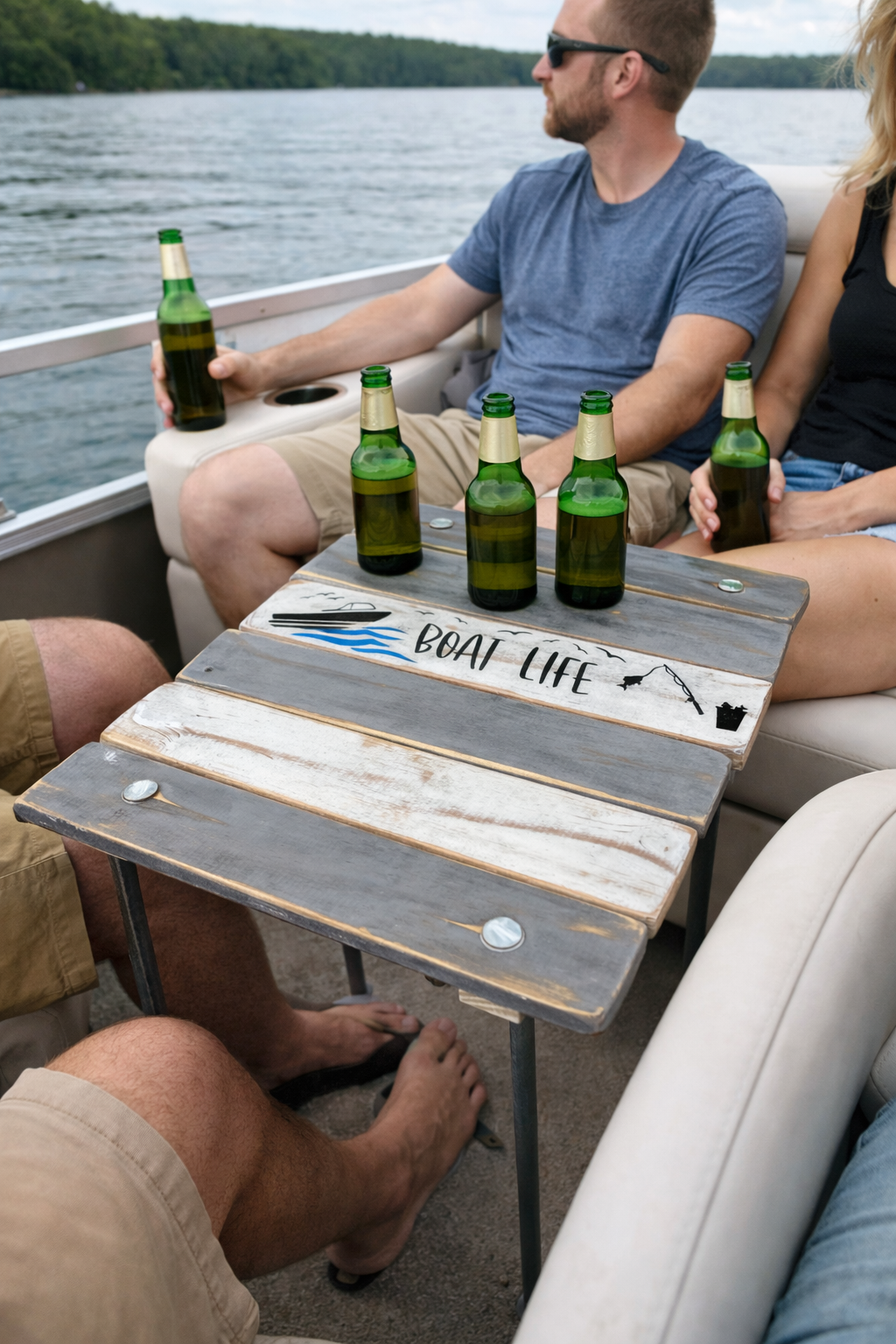 People sitting on a boat with beer bottles, a wooden table with a painted sign that says 'Boat Life', and a water body and trees in the background.