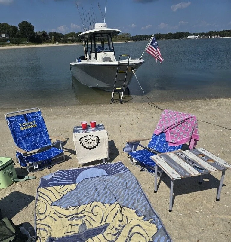 Portable cedar roll-out table set up on sandy beach with chairs, drinks, and anchored boat in background