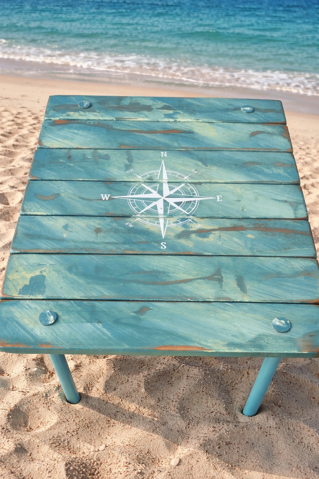 turquoise portable beach table with a white compass rose on the beach with small waves in background